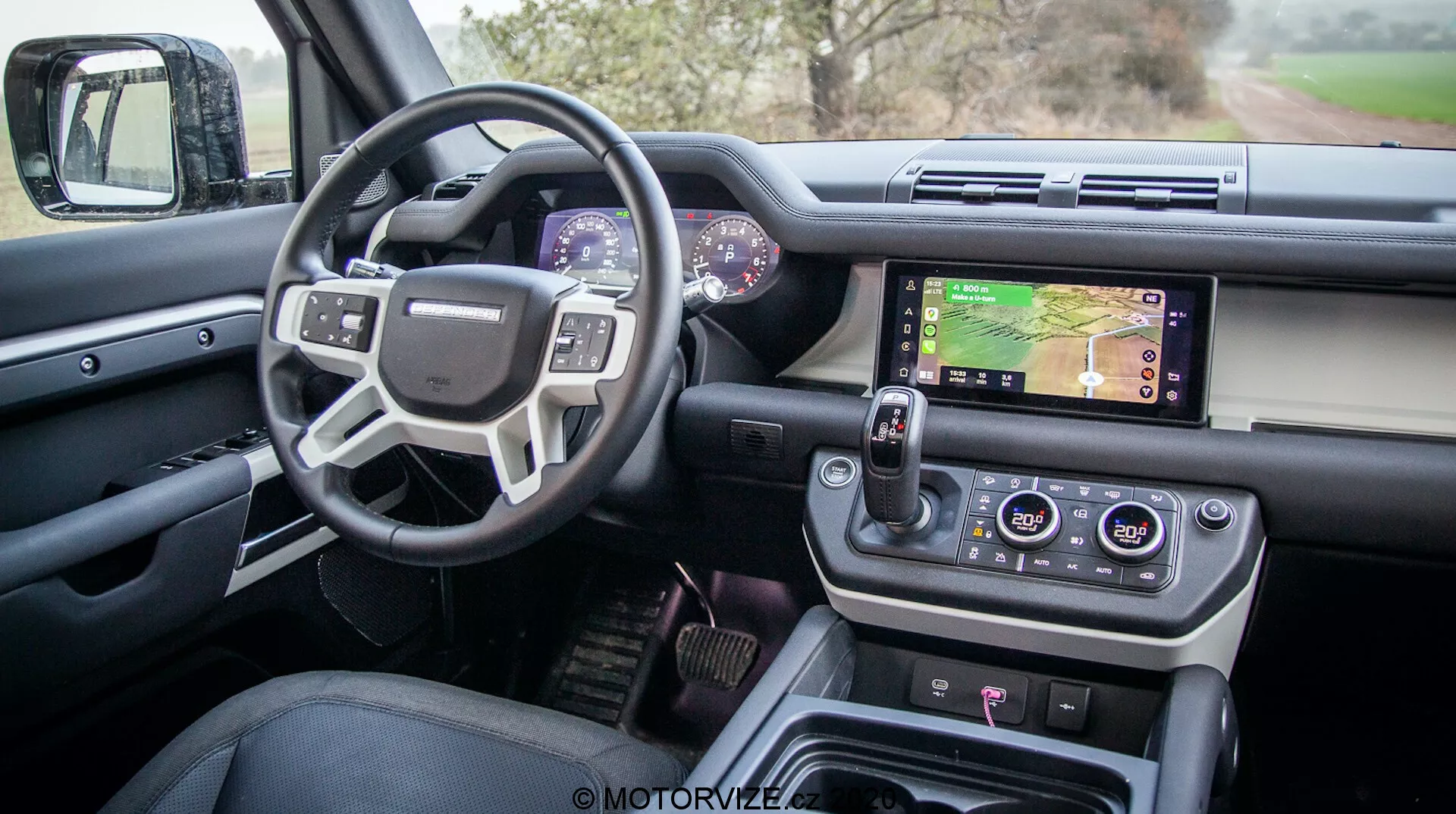 Vista interna di una moderna Land Rover Defender 2020 dalla porta del conducente che mostra il design sofisticato e le caratteristiche avanzate incluse una porta del conducente aperta con comandi per finestrini e specchietti, un volante multifunzionale con pulsanti di controllo, un quadro strumenti digitale, un sistema di infotainment centrale con touchscreen che mostra una mappa, manopole per il controllo del clima, un moderno selettore del cambio rotativo e pulsanti di controllo del terreno, il tutto in una finitura di lusso e alta qualità.
