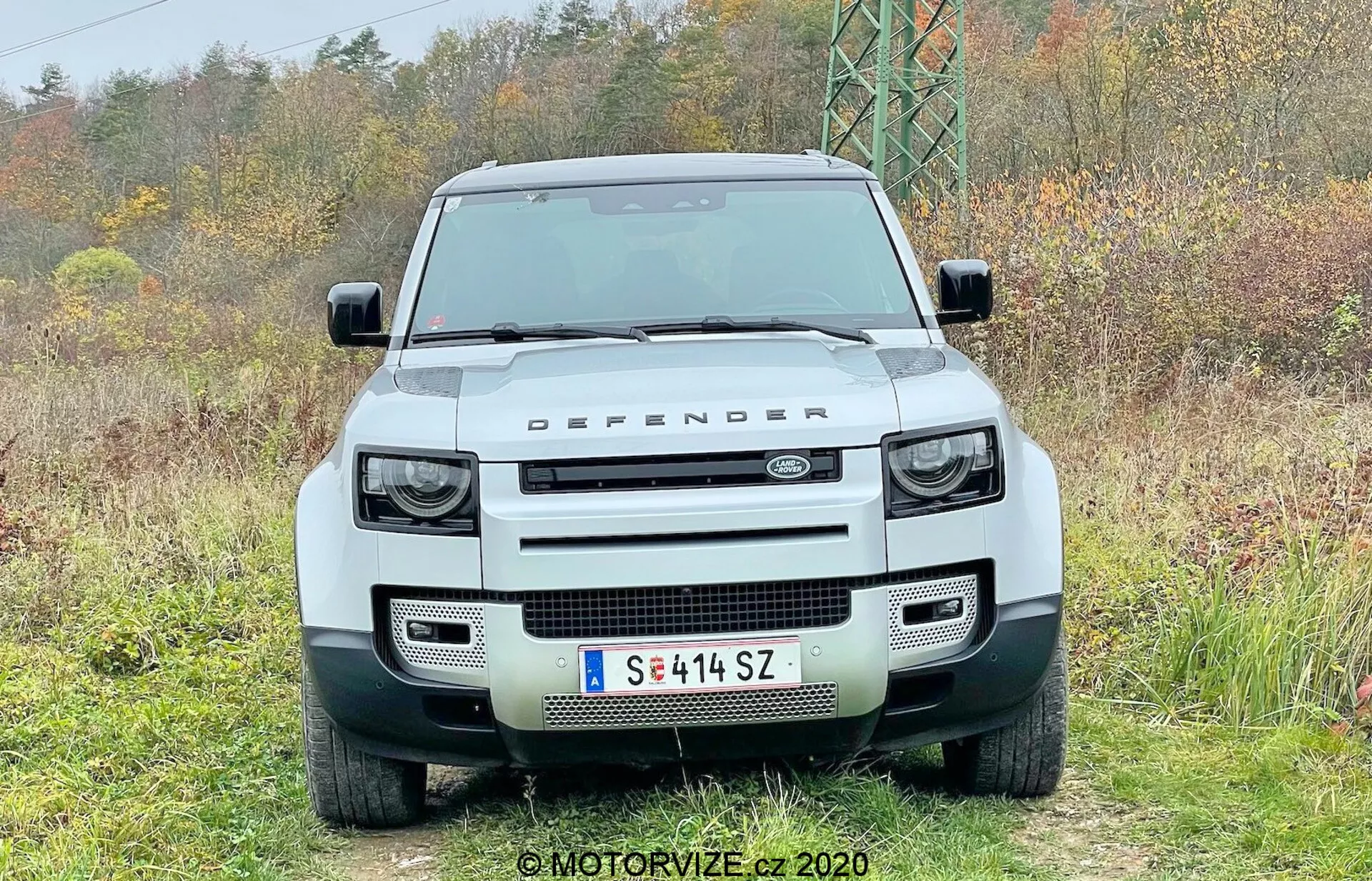 Vista frontale di un Land Rover Defender 2020 bianco 4x4, con una griglia rettangolare prominente affiancata da fari rotondi a LED con luci diurne quadrate integrate, robusto paraurti anteriore con fessure rettangolari, cofano piatto con la scritta 'LAND ROVER' e il marchio 'DEFENDER' sul bordo anteriore, parabrezza piatto, targa europea montata centralmente e sensori di parcheggio, catturato fermo in un ambiente naturale.