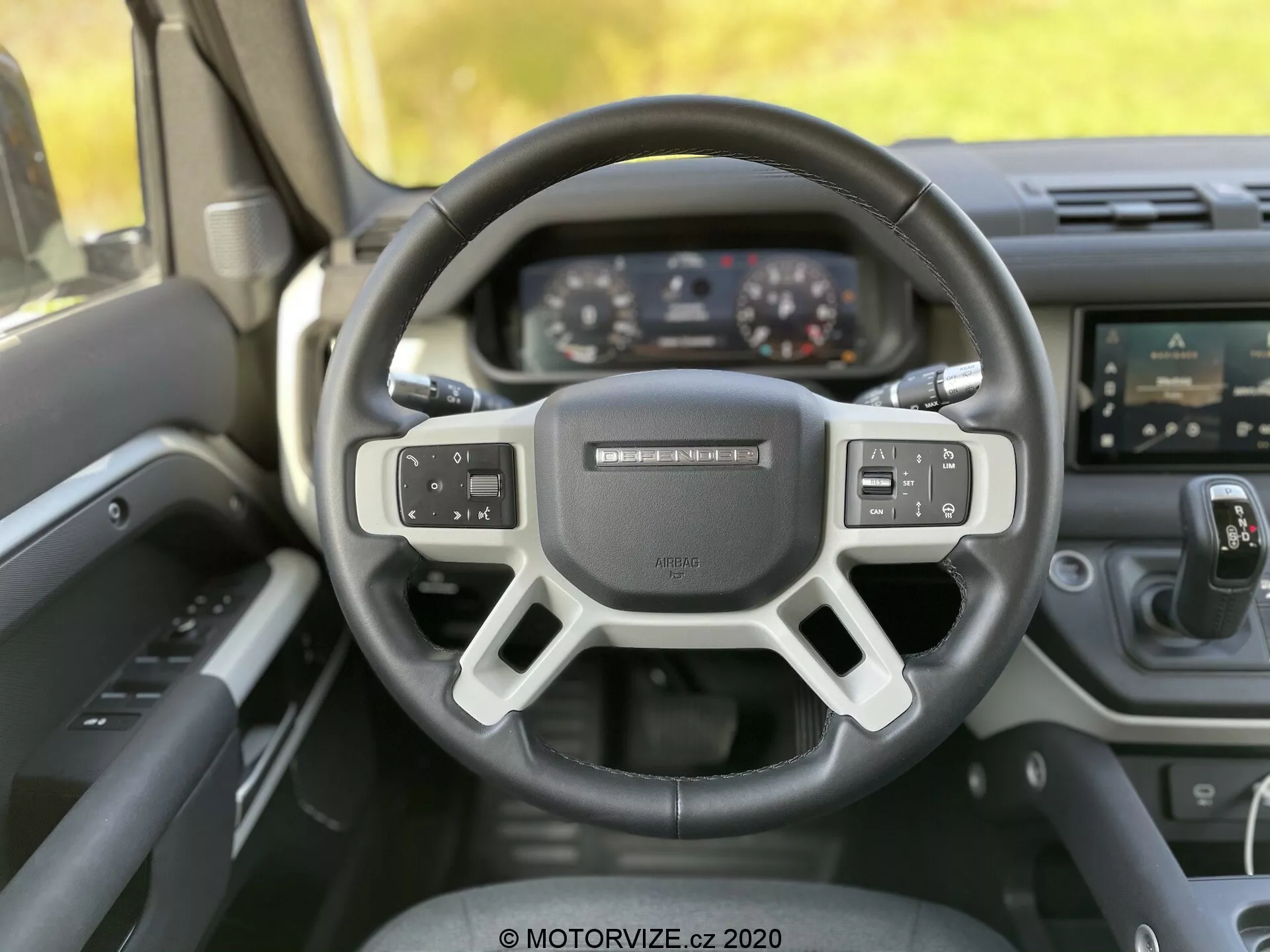 Vista interna del Land-Rover Defender 2020 dalla prospettiva del conducente, con il volante moderno a tre razze con comandi integrati per audio, comandi vocali e impostazioni del cruise control. Dietro il volante, il quadro strumenti presenta display digitali per il tachimetro, il contachilometri e un display multifunzionale. L'immagine cattura anche la console centrale con un sistema di infotainment touchscreen e pulsanti per il controllo del clima sotto. L'angolo della foto è leggermente inclinato verso l'alto, evidenziando il design ergonomico, la tecnologia avanzata e i materiali di alta qualità del veicolo fermo.
