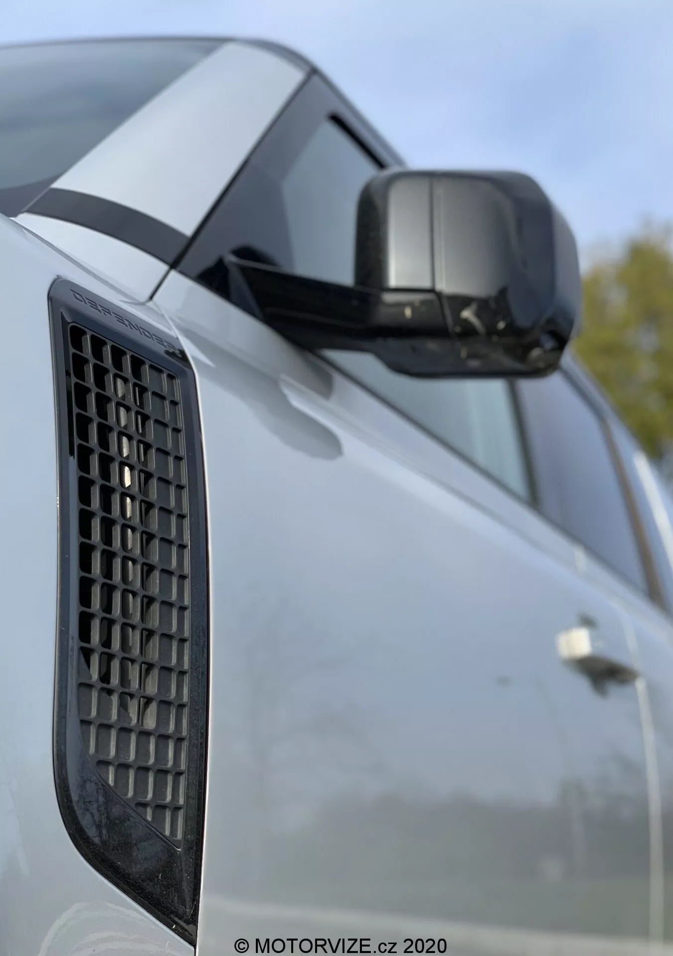 Primo piano del Land Rover Defender 2020 che evidenzia il parafango laterale con griglia, lo specchietto laterale con indicatore di direzione integrato e una parte della porta con linee caratteristiche marcate; la foto mette in risalto gli elementi di design del veicolo con uno sfondo sfocato che suggerisce un ambiente esterno.