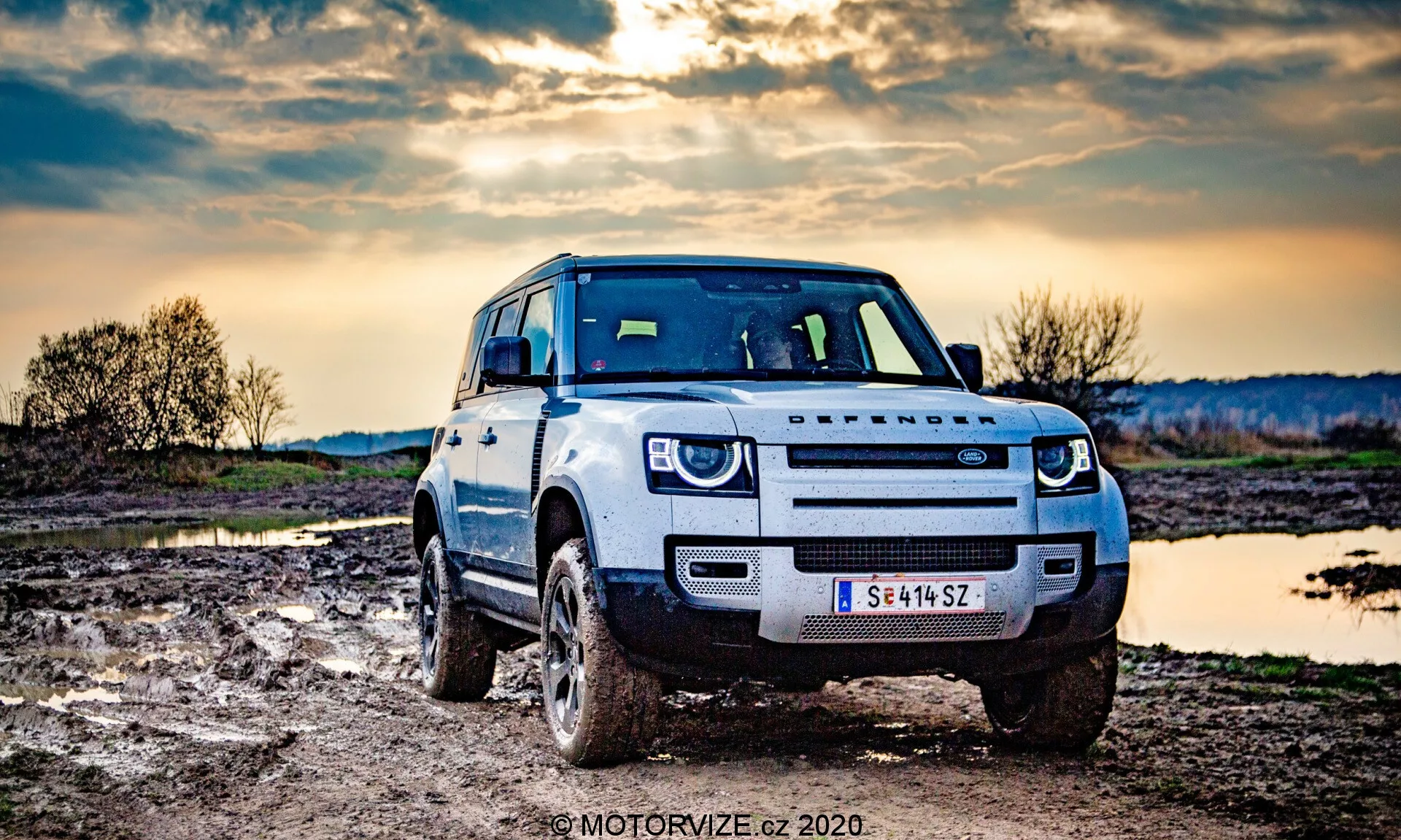 Fotografia di un SUV Land Rover Defender ripresa da un angolo anteriore, leggermente fuori centro, che evidenzia la parte anteriore del veicolo e le sue capacità fuoristrada. Il veicolo è di colore chiaro, con sporco sulle gomme e sul pannello inferiore che indica che è stato guidato in fuoristrada. L'auto ha uno snorkel per guadi profondi e fari a LED. L'ambiente circostante rurale presenta un terreno fangoso e un corpo d'acqua ferma, con un drammatico cielo serale sullo sfondo.
