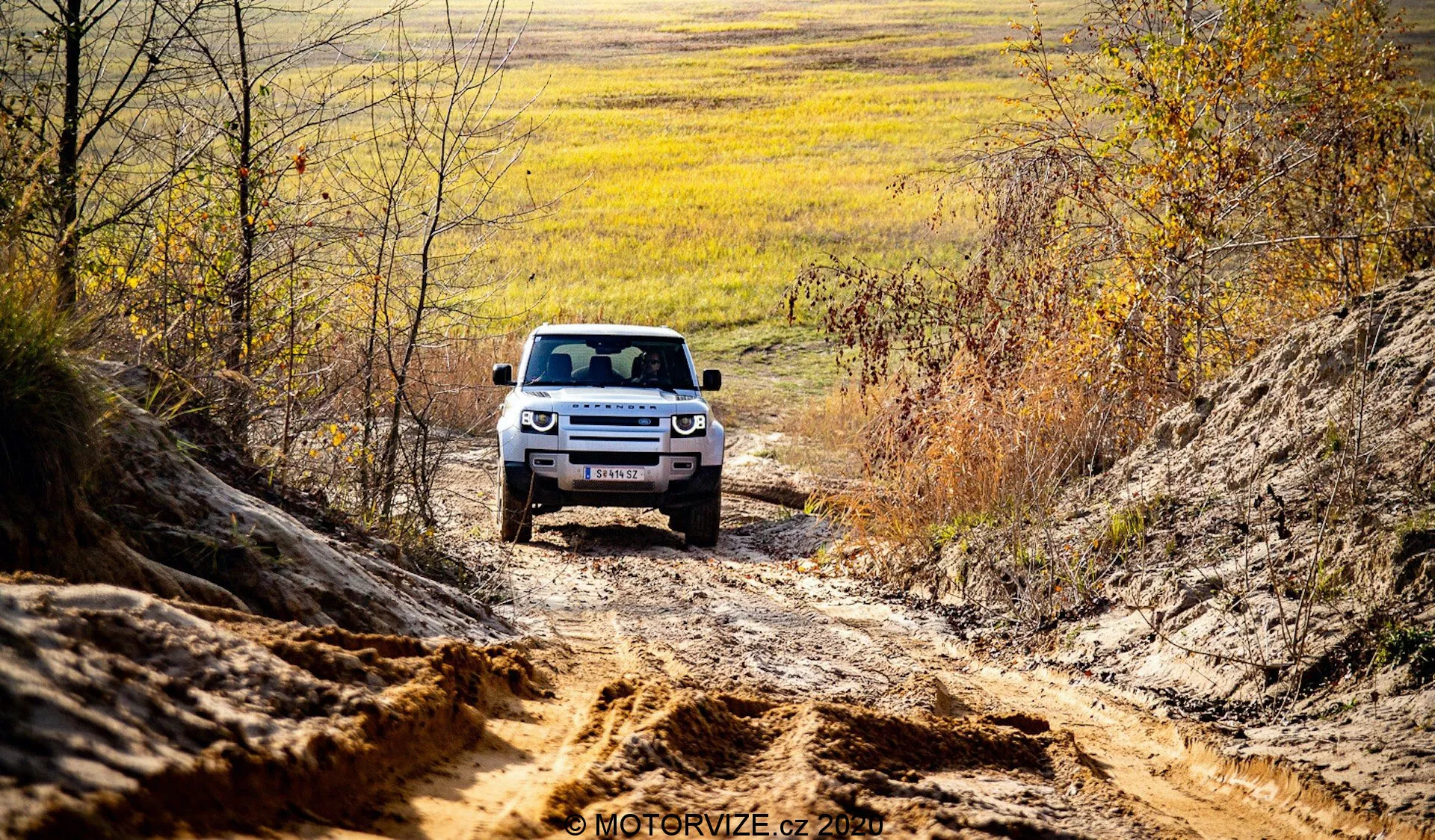 Un SUV Land Rover Defender 2020 è visto frontalmente durante la guida fuoristrada, posizionato centralmente contro uno sfondo di terreno accidentato e ambiente naturale. La parte anteriore visibile del veicolo mostra un'avanzata articolazione delle sospensioni, fari a LED e una griglia caratteristica con listelli orizzontali. Catturando il SUV in un ambiente rurale con tonalità autunnali e luce naturale, la prospettiva esalta la robusta e avventurosa presenza del veicolo.