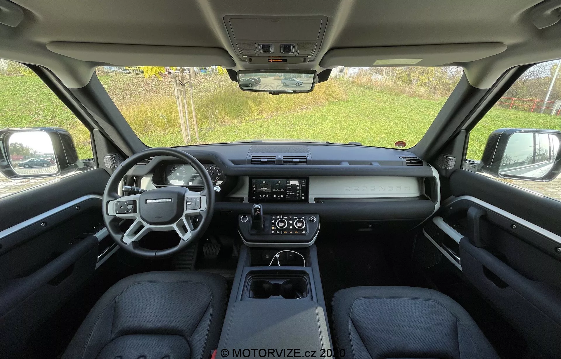 Vista interna di una Land Rover Defender 2020 dal sedile posteriore, che mostra un abitacolo elegante e moderno con volante multifunzione, cruscotto orizzontale con display digitale centrale, sistema di infotainment touch screen prominente, controllo del clima a doppia zona e console centrale pulita con selettore del cambio elettronico e portabicchieri.