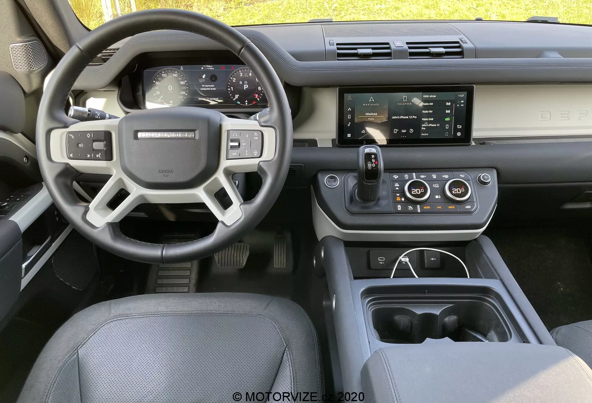 Scatto dell'interno di una Land Rover Defender 2020 dal sedile posteriore centrale, che mostra il cruscotto, il volante con pulsanti di controllo, il quadro strumenti totalmente digitale e il display centrale di infotainment con tecnologie intelligenti e di comfort.