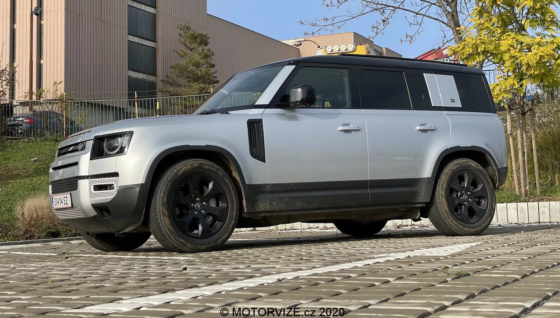 Fotografia di un moderno SUV Land Rover Defender 2020 scattata da un'angolazione bassa per accentuare la sua posa robusta e imponente. Il veicolo è mostrato in una vista di tre quarti dalla parte anteriore e laterale, evidenziando la sua griglia distintiva, fari a LED, paraurti con luci fendinebbia e sensori, cerchi in lega nera con design a più razze e pneumatici a profilo basso, specchietti laterali neri con tecnologie aggiuntive potenziali, vetri oscurati, linea del tetto prominente, maniglie delle portiere a filo, design forte dell'arco ruota e colore argento chiaro o grigio contrastato con elementi di rifinitura neri. Il SUV è parcheggiato su un pavimento in ciottoli con un edificio e vegetazione sullo sfondo in condizioni di luce diurna.