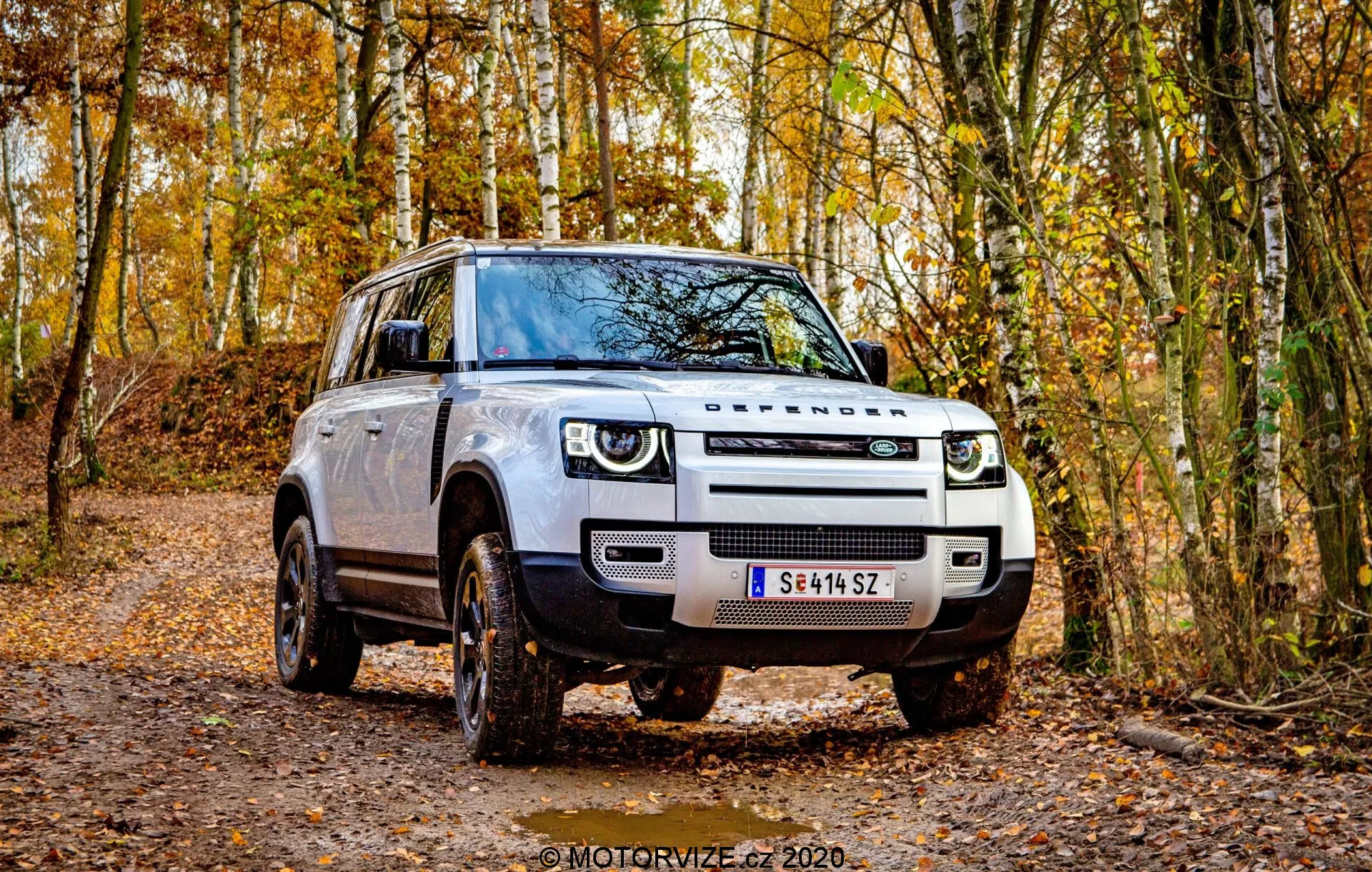 Questa immagine presenta un SUV Land Rover Defender 2020 posizionato su un sentiero forestale non asfaltato, che mostra un design moderno e squadrato da un angolo frontale tre quarti. Sono chiaramente visibili la griglia, i fari e il profilo laterale del veicolo. I fari LED distintivi con luci diurne, la griglia prominente e i fendinebbia pronunciati sono evidenziati. Le grandi ruote in lega con battistrada fuoristrada, gli specchietti laterali con indicatori di direzione integrati e l'altezza da terra elevata enfatizzano la sua capacità fuoristrada. L'esterno robusto, le maniglie delle porte tradizionali e il colore neutro della carrozzeria migliorano ulteriormente il suo fascino come un SUV moderno pronto per l'avventura.