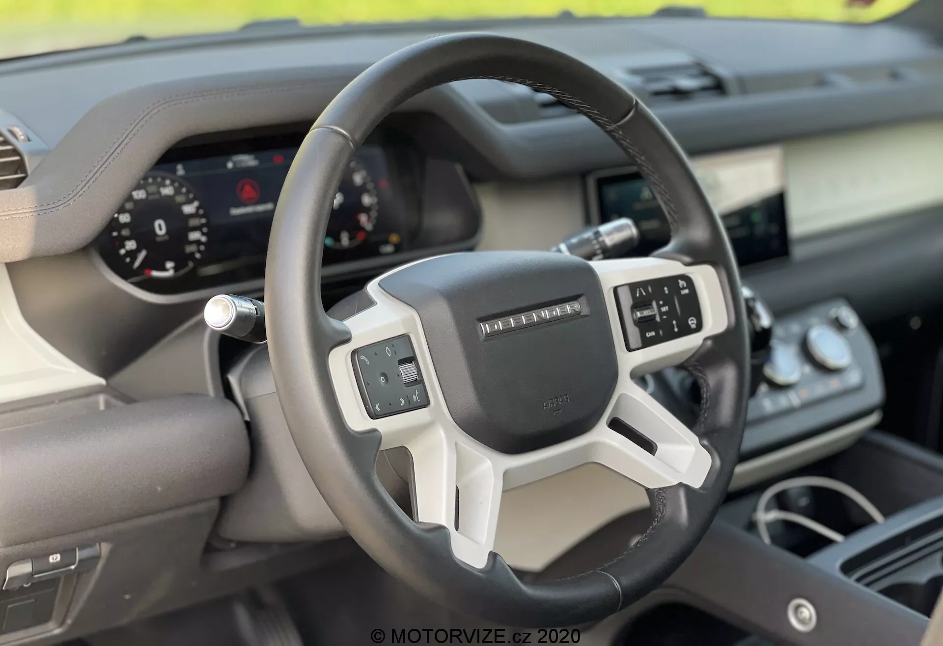 Vista interna del Land Rover Defender 2020 focalizzata sul volante e sul cruscotto dalla prospettiva del sedile anteriore del passeggero. Il volante moderno a tre razze presenta controlli multifunzione e un quadro strumenti digitale per una visualizzazione chiara degli indicatori. La combinazione di materiali come pelle e finiture metalliche o plastiche sottolinea il design ergonomico e tecnologico del veicolo.