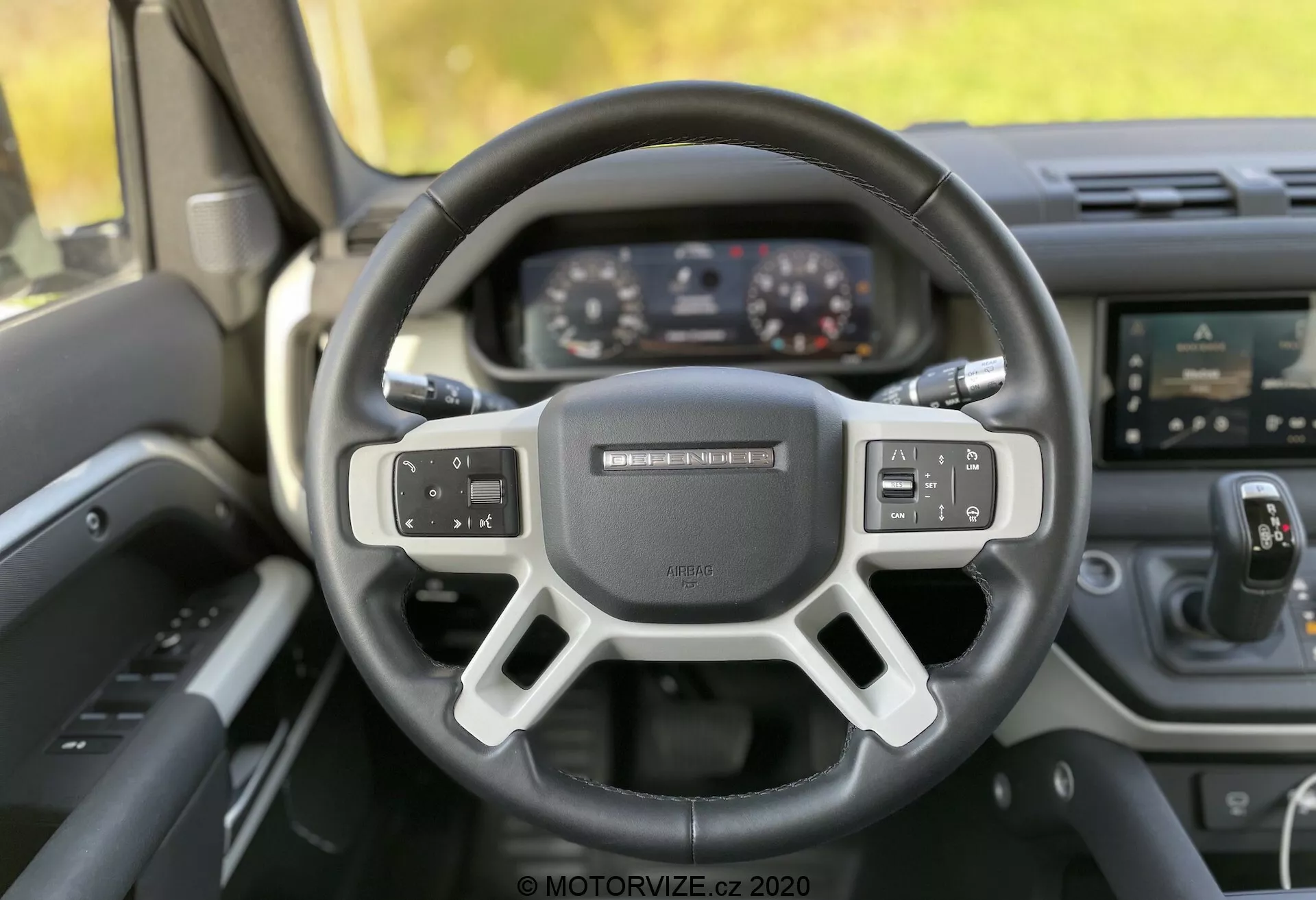 Vista interna della Land Rover Defender 2020, che mostra la prospettiva del guidatore con particolare attenzione al volante e al cruscotto. Il volante a tre razze con fondo piatto presenta comandi multifunzionali e un badge sfocato al centro. Dietro di esso, un pannello strumenti digitale mostra la velocità e altre informazioni, indicativo della tecnologia moderna. La console centrale mostra un sistema di infotainment touch-screen con varie icone e pulsanti fisici per il controllo del clima e dell'audio. Gli interni sono realizzati in materiale resistente, probabilmente plastica dura o fibra di carbonio. L'immagine di alta qualità, scattata con luce naturale, utilizza un effetto bokeh per evidenziare il volante e i controlli.