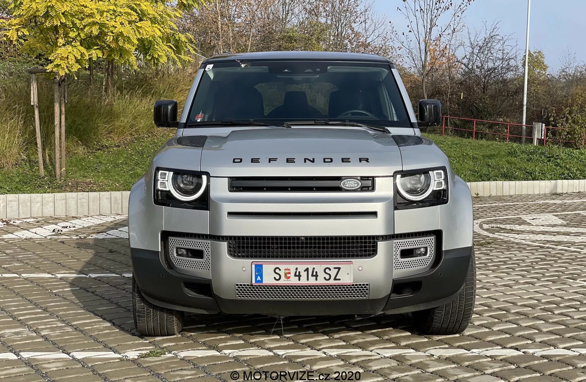 Vista frontale del SUV Land Rover Defender 2020, che mostra la sua griglia distintiva, i fari e il cofano con la scritta 'DEFENDER'. L'immagine, centrata e scattata da un'angolazione leggermente elevata, mette in risalto i fari a LED moderni, il motivo geometrico della griglia, i sensori della telecamera frontale, i sensori di parcheggio, la piastra di scorrimento anteriore e i punti di traino. Il veicolo è posizionato contro uno sfondo esterno semplice con cespugli e una ringhiera, sotto la luce naturale del giorno.