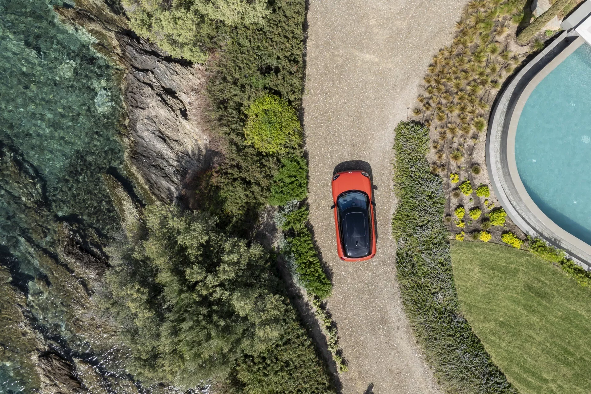Veduta aerea della Mini Aceman 2024 (J05) che mostra il suo tetto, cofano, baule e parte del profilo laterale parcheggiata vicino a una scogliera e a un corpo d'acqua, circondata da una lussureggiante vegetazione, evidenziando la caratteristica del tetto solare dell'auto