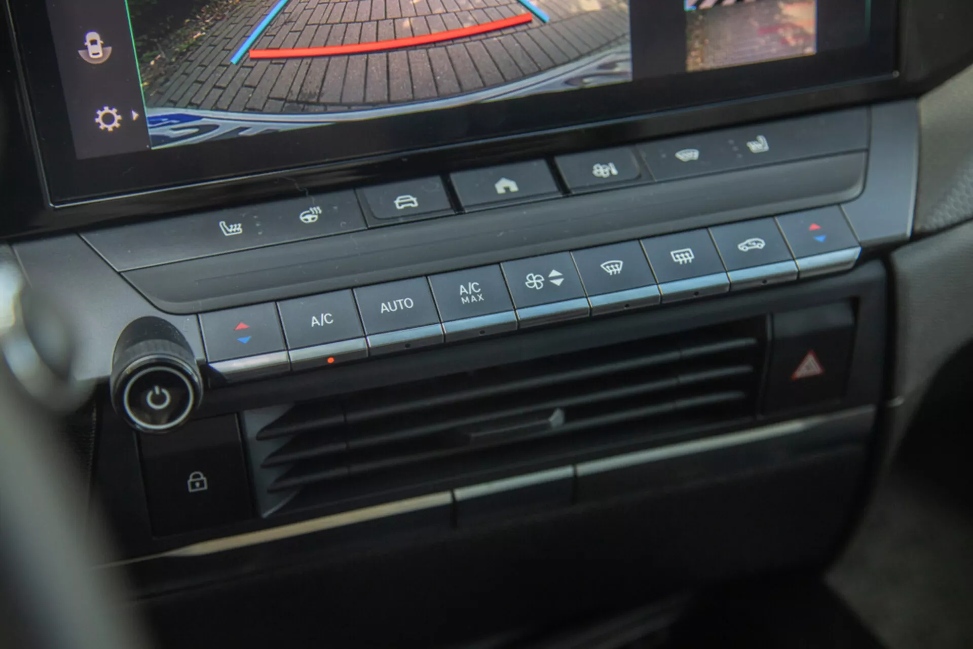 Console centrale e pannello di controllo climatico dell'Opel Astra 2022, evidenziando il pomello di regolazione della temperatura, la velocità della ventola, la distribuzione dell'aria e i pulsanti di controllo clima automatico con display digitale che mostra la telecamera posteriore