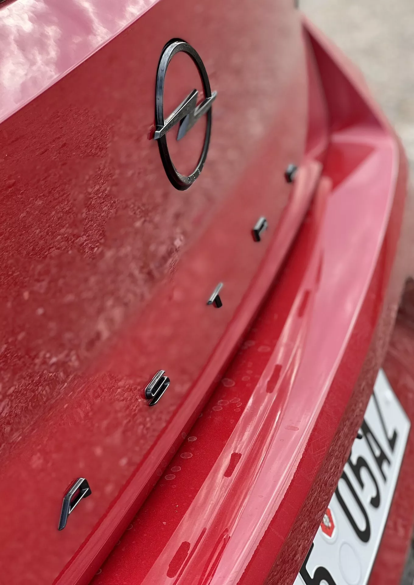 Veduta ravvicinata della sezione posteriore dell'Opel Astra 2022 con vernice rossa e strato di polvere visibile, includendo l'emblema del produttore e sensori per l'assistenza al parcheggio