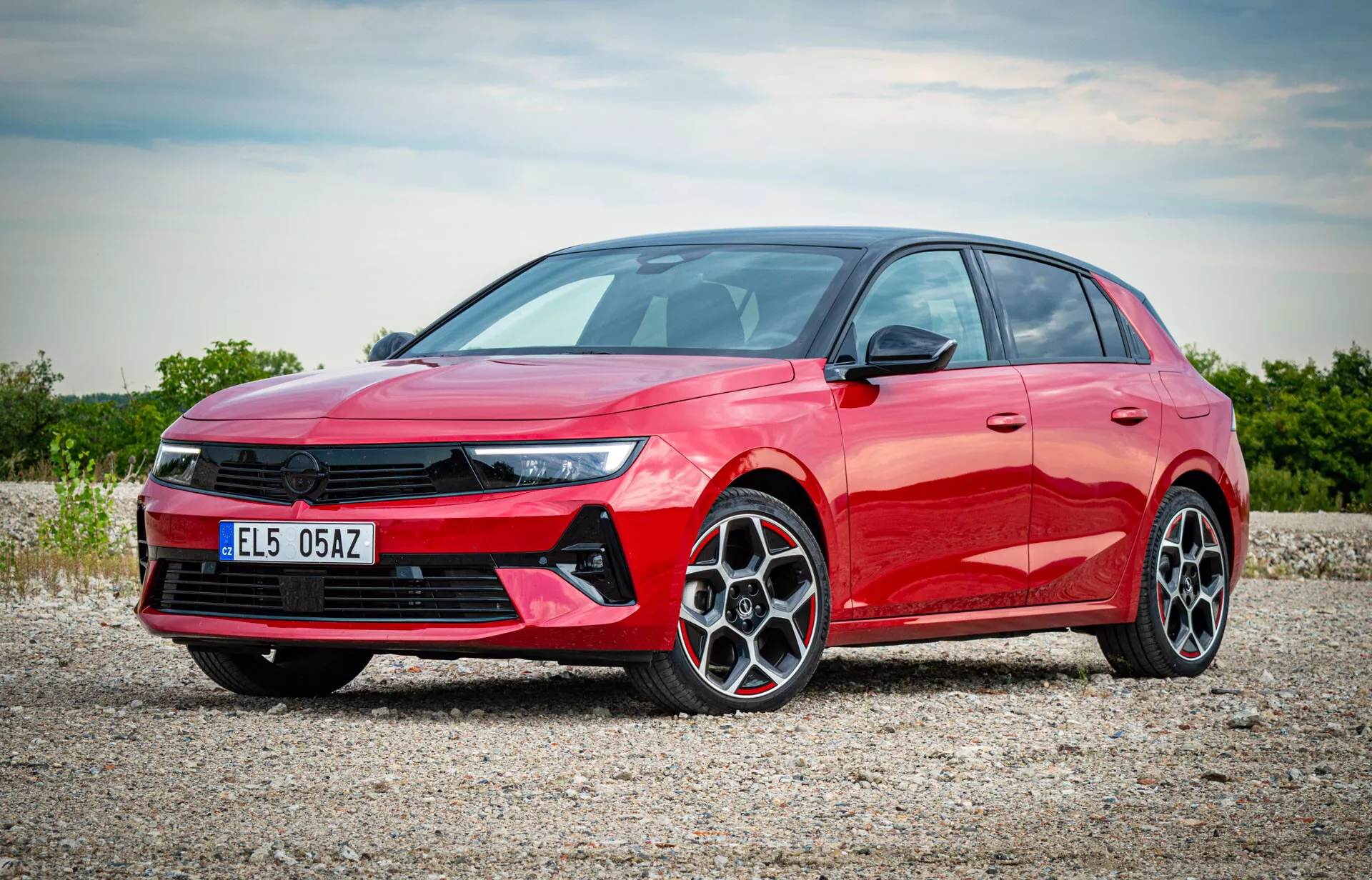 Opel Astra hatchback rosso 2022 fotografato da una vista frontale a tre quarti che mostra i fari LED, la griglia anteriore prominente, le ruote in lega a più razze e il profilo laterale elegante su una superficie di ghiaia con luce solare diffusa che illumina le caratteristiche moderne dell'auto