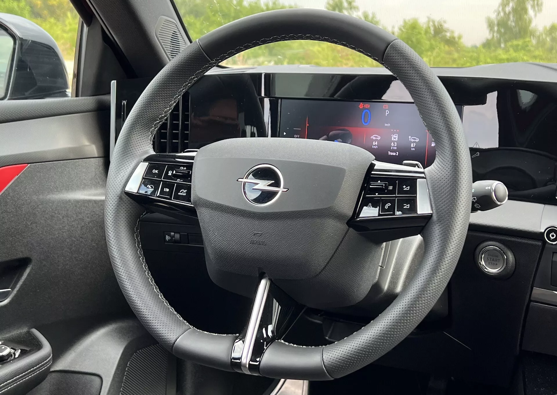 Vista interna dettagliata del volante dell'Opel Astra 2022 con design a tre razze con comandi integrati e un display del cruscotto completamente digitale dietro, evidenziando la moderna tecnologia e l'ergonomia del veicolo