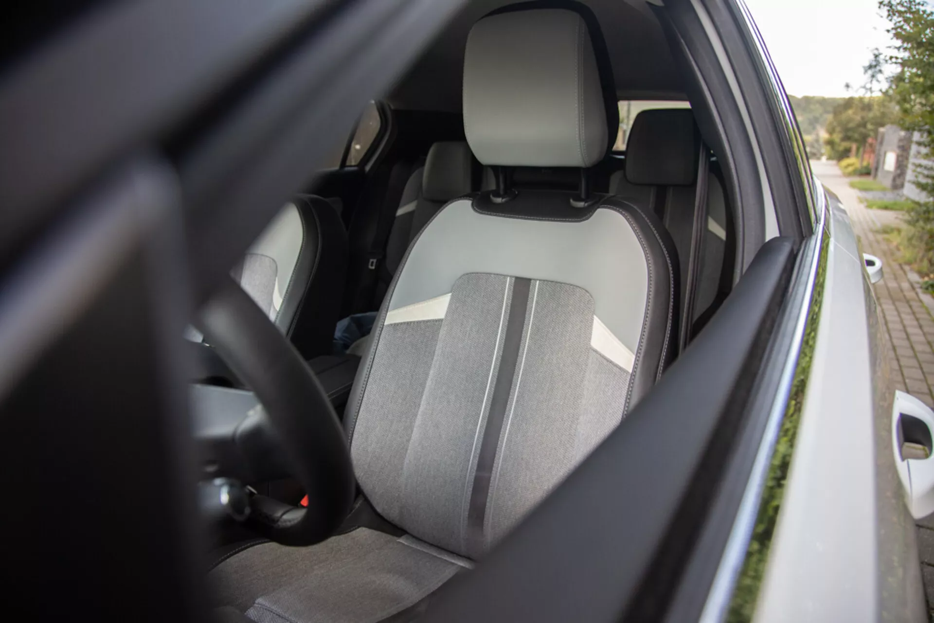 Vista ravvicinata dell'interno lato guidatore della Opel Astra 2022 da un angolo laterale esterno che mostra il sedile del guidatore in tessuto bicolore, poggiatesta integrato, parte del volante con comandi, accenno del cruscotto, cintura di sicurezza inserita, interno della porta con potenziali comandi per finestrini elettrici e alloggiamento dello specchio laterale del colore della carrozzeria, con focus su ergonomia, sicurezza e funzionalità.