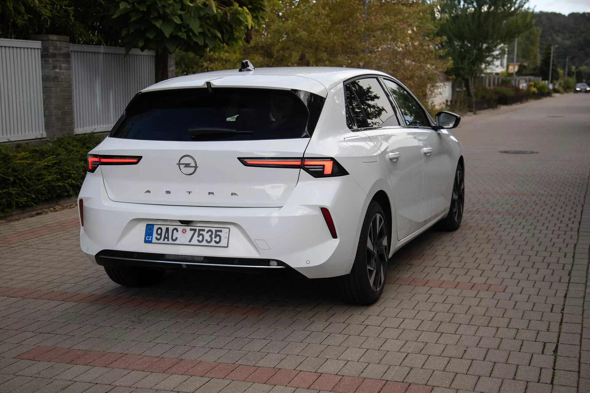 Berlina moderna Opel Astra 2022 in bianco, parcheggiata su una superficie di mattoni incastro, mostrando un angolo posteriore di tre quarti con un focus su fanali posteriori LED ad alta tecnologia, telecamera posteriore, sensori di parcheggio, profilo laterale parziale incluso la ruota e la porta posteriore destra, collegati da una barra luminosa orizzontale attraverso il coperchio del bagagliaio, dotata di antenna a pinna di squalo per una connettività migliorata, adatto per illustrare il design posteriore del veicolo e la tecnologia automobilistica moderna