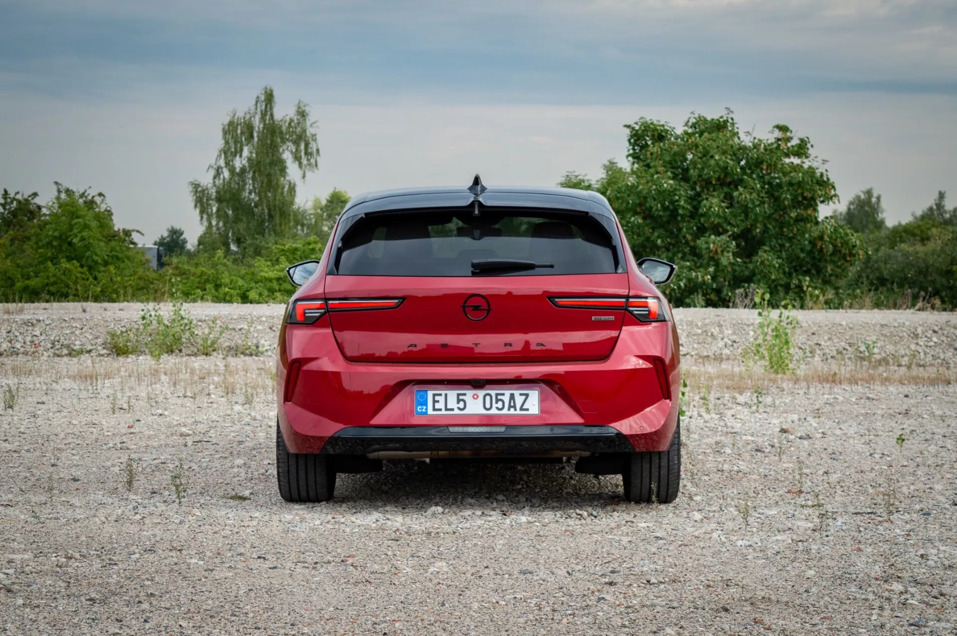 Opel Astra 2022 fotografata da dietro su una superficie di ghiaia che mette in mostra fanali posteriori con una barra luminosa di connessione, sensori di parcheggio e l'assenza di porte di scarico indicando un possibile modello elettrico o ibrido, su uno sfondo di vegetazione e cielo leggermente nuvoloso.
