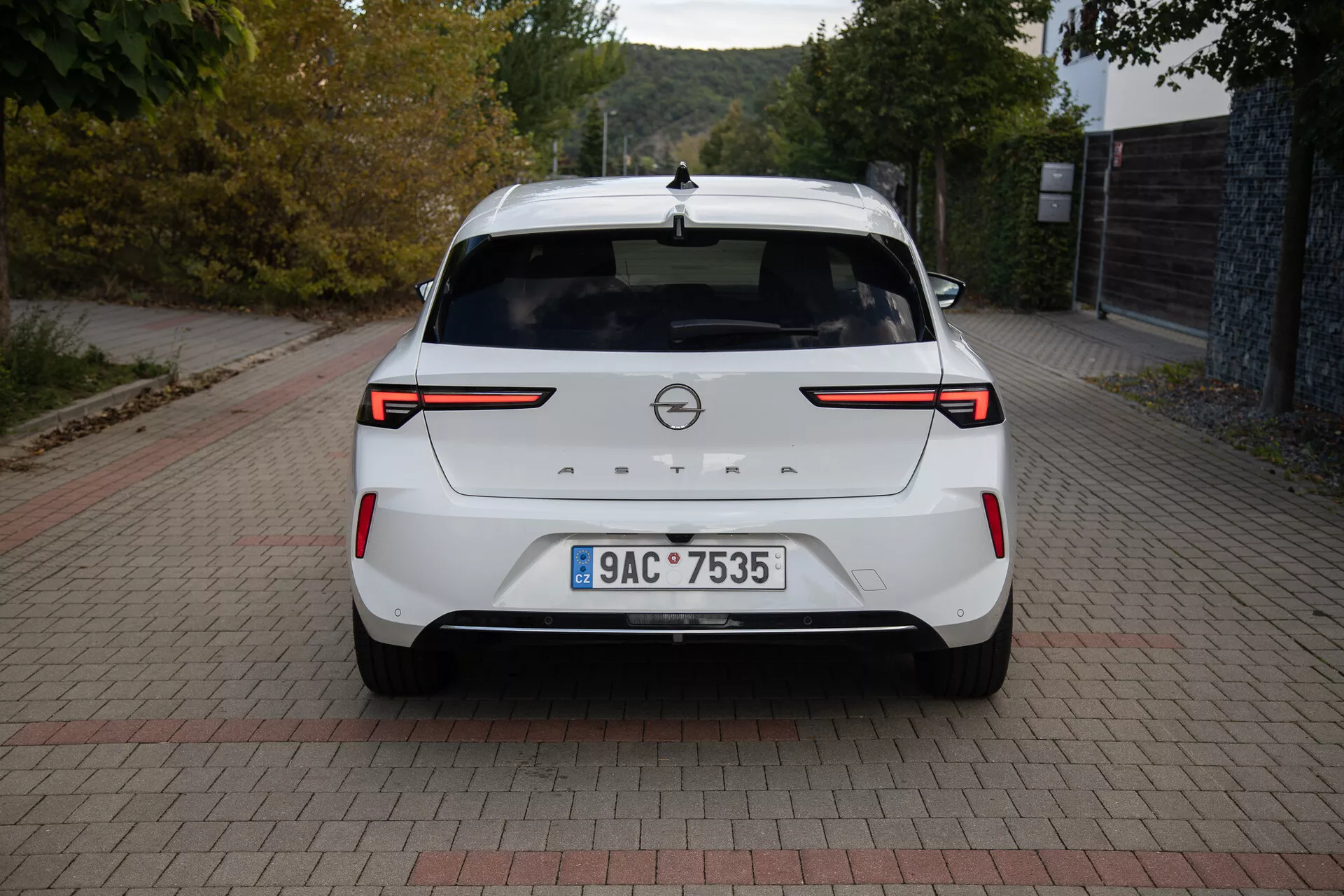 Vista posteriore di una Opel Astra 2022 bianca che mostra fari posteriori LED, sensori / telecamera di parcheggio posteriori, badge del marchio, targa, spoiler posteriore, luce di stop montata in alto, antenna a pinna di squalo e una finitura della vernice pulita, parcheggiata su una superficie a motivo di mattoni alla luce del giorno.