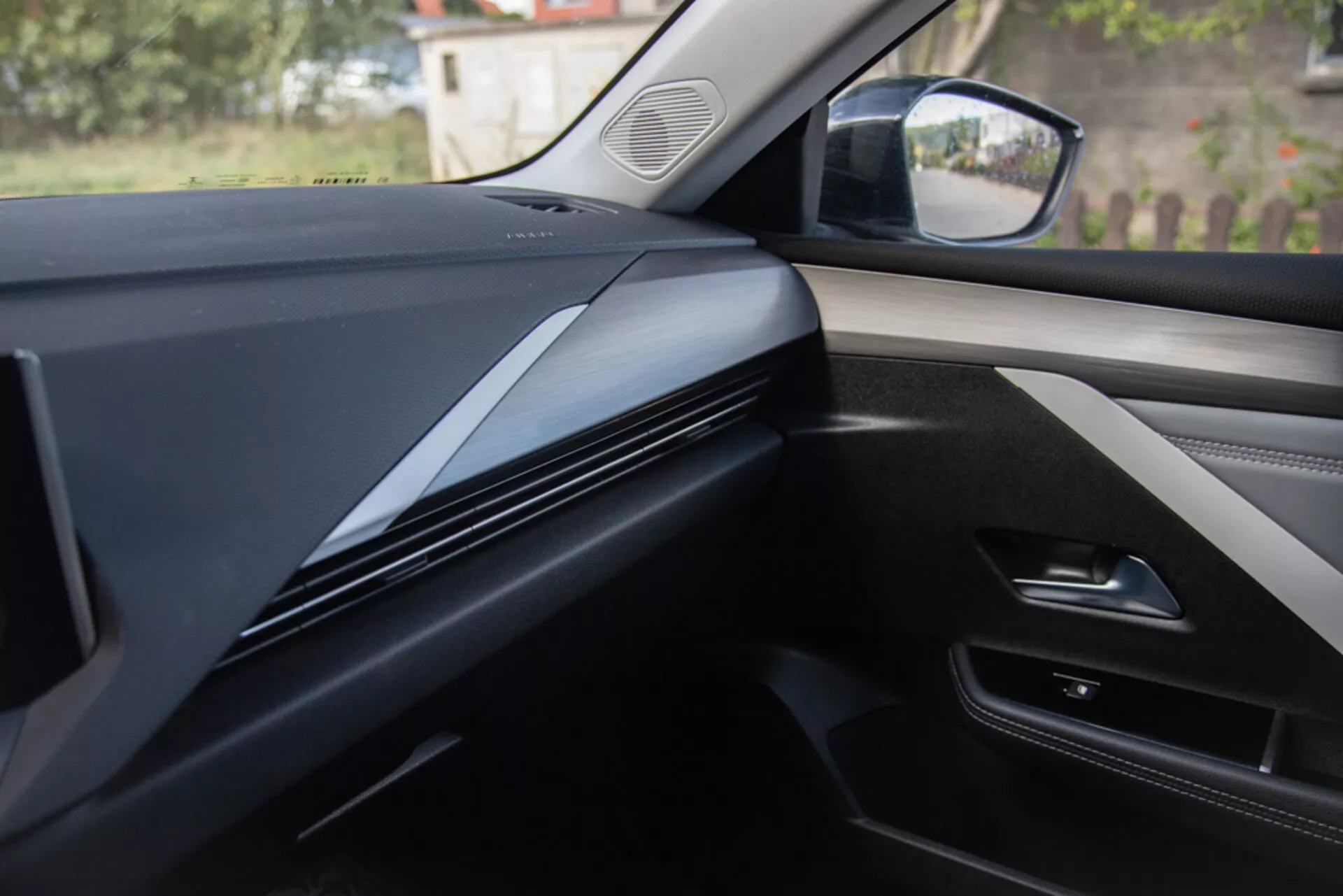 Vista interna dalla prospettiva del sedile passeggero che mostra il design del cruscotto, rifiniture e pannello della porta di una Opel Astra 2022, con linee contornate scure ed eleganti con un focus sulla texture dei materiali e potenziali integrazioni tecnologiche
