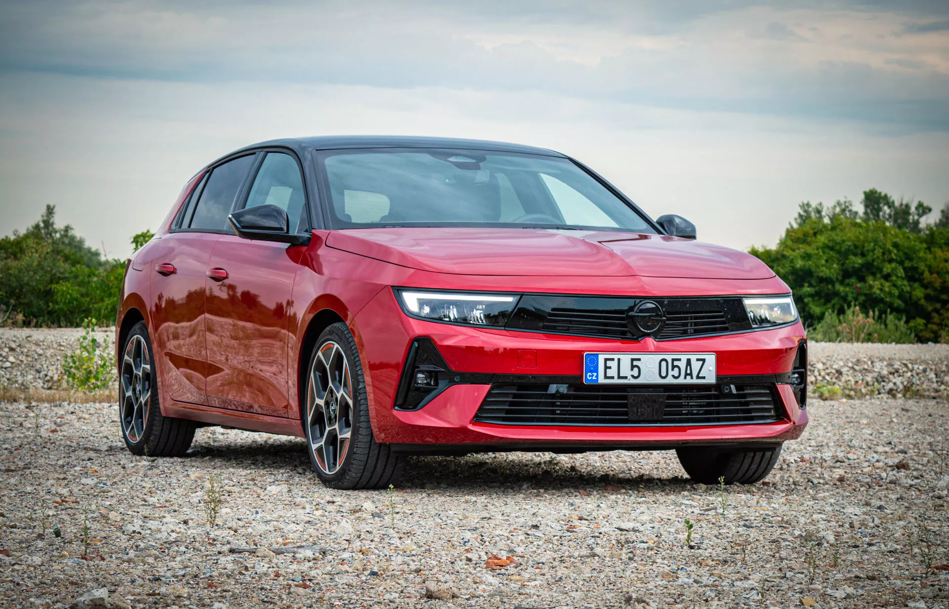 Opel Astra 2022 in rosso vibrante, mostra un design moderno con fari LED angolari, griglia frontale prominente, paraurti anteriore sportivo e cerchi in lega a più razze, esposta in un ambiente naturale all'aperto da una vista frontale di tre quarti che evidenzia il profilo aerodinamico e le caratteristiche premium dell'auto