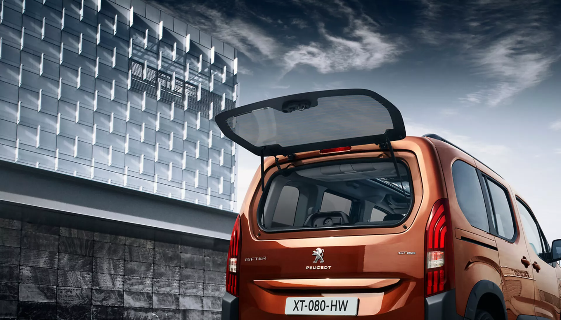 Vista angolare posteriore della Peugeot Rifter 2018 che mostra il suo design hatchback, finitura metallica, portellone aperto con spazio di carico visibile, luci posteriori LED e sfondo urbano.