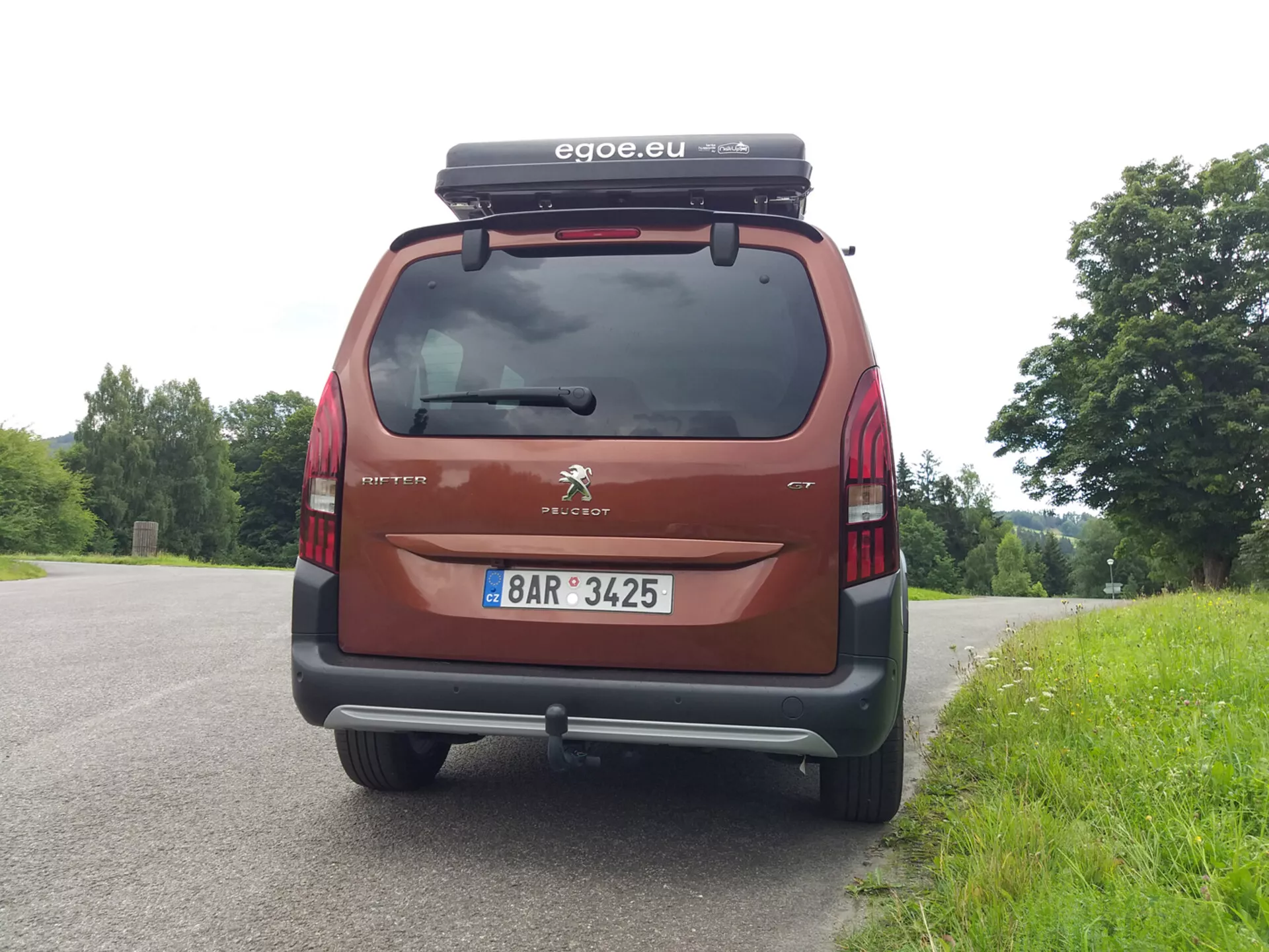 Vista posteriore di una Peugeot Rifter 2018, mostrando il lunotto posteriore, le luci di coda, il paraurti e la targa, con il logo della Peugeot e il nome del modello Partner visibili. L'auto, parcheggiata su uno sfondo naturale, è dotata di una scatola da tetto e presenta un accessorio marchiato 'egoe.eu', in colore rosso/marrone.