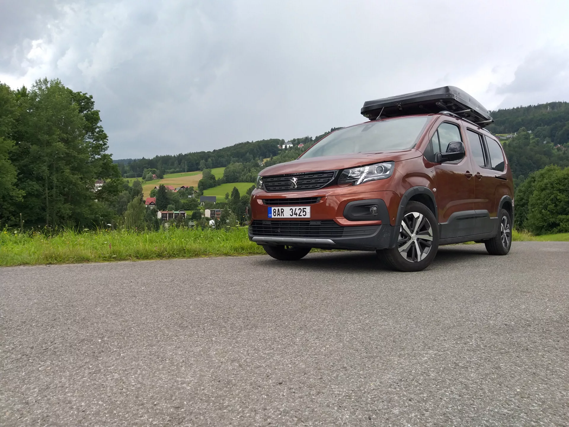 SUV Peugeot Rifter 2018 con vernice metallica e box sul tetto catturata da un angolo basso che mette in evidenza la griglia, il paraurti e le ruote in lega, mostrando capacità off-road e design moderno inclusi fari LED e sistemi ADAS, parcheggiata su una superficie pavimentata contro un paesaggio rurale