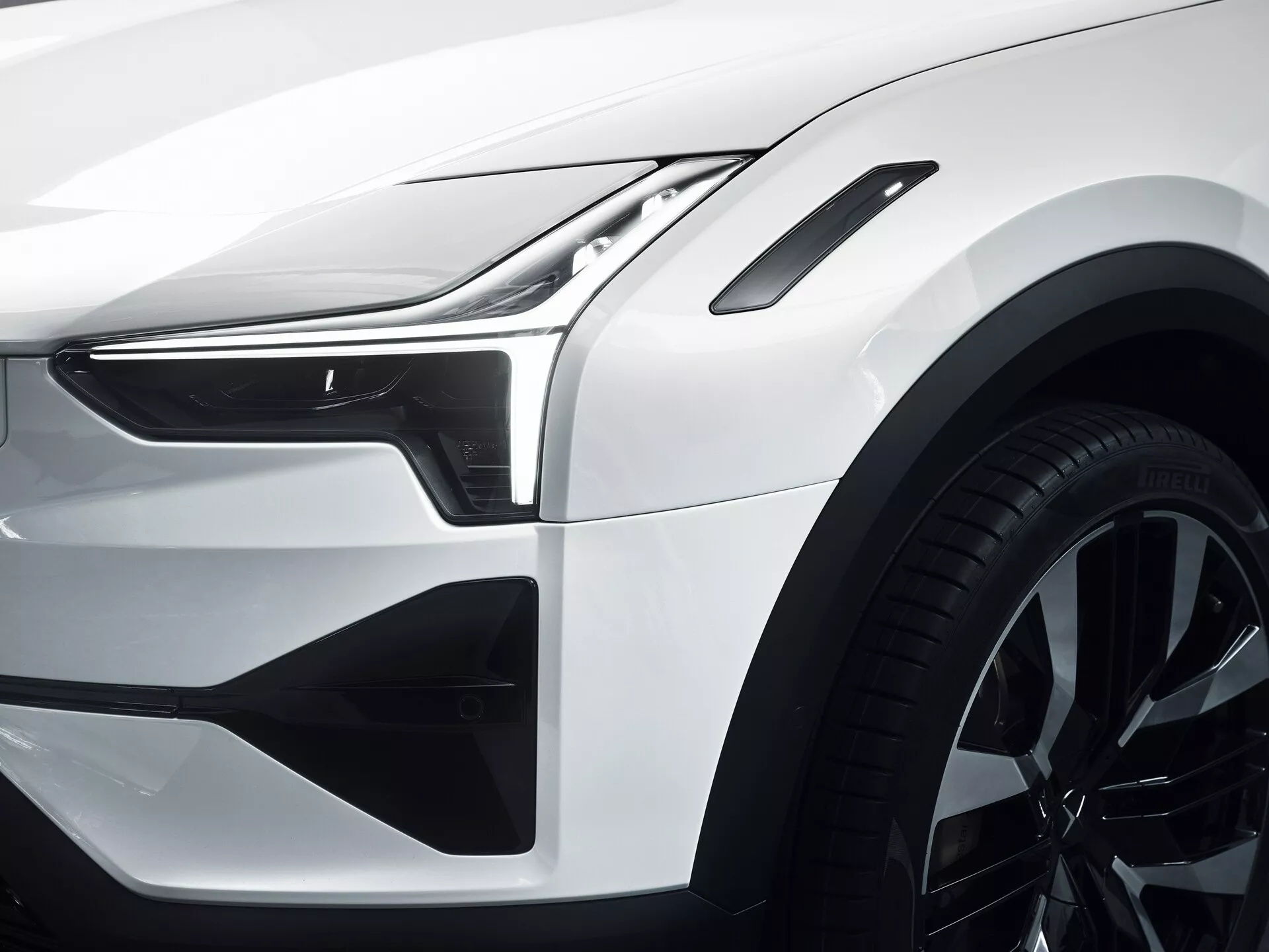 Vista ravvicinata del quarto anteriore del Polestar 3 2023 con faro LED e luci diurne, contorni del cofano e del parafango aerodinamici, prese d'aria del paraurti anteriore stilizzate, ruota in lega a più razze, distanze tra i pannelli precisi e linee della carrozzeria, finitura bianca lucida, indicando un design e una tecnologia automobilistica avanzata