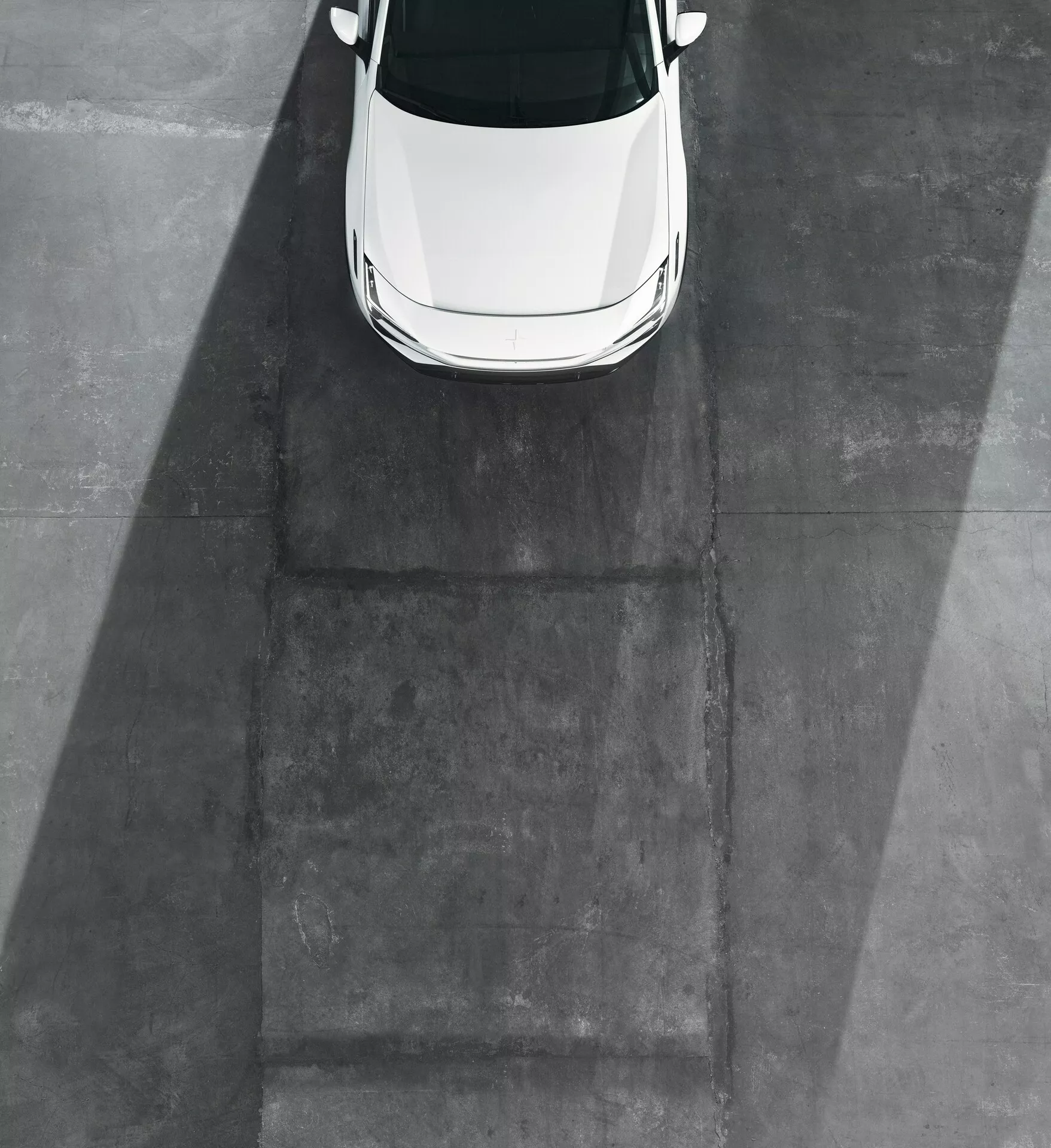 Modello Polestar 3 2023 bianco catturato in vista dall'alto evidenziando il tetto, il cofano e parte del parabrezza anteriore contro una superficie in cemento grigio senza ruote o lati visibili, indicando luce mattutina o pomeridiana