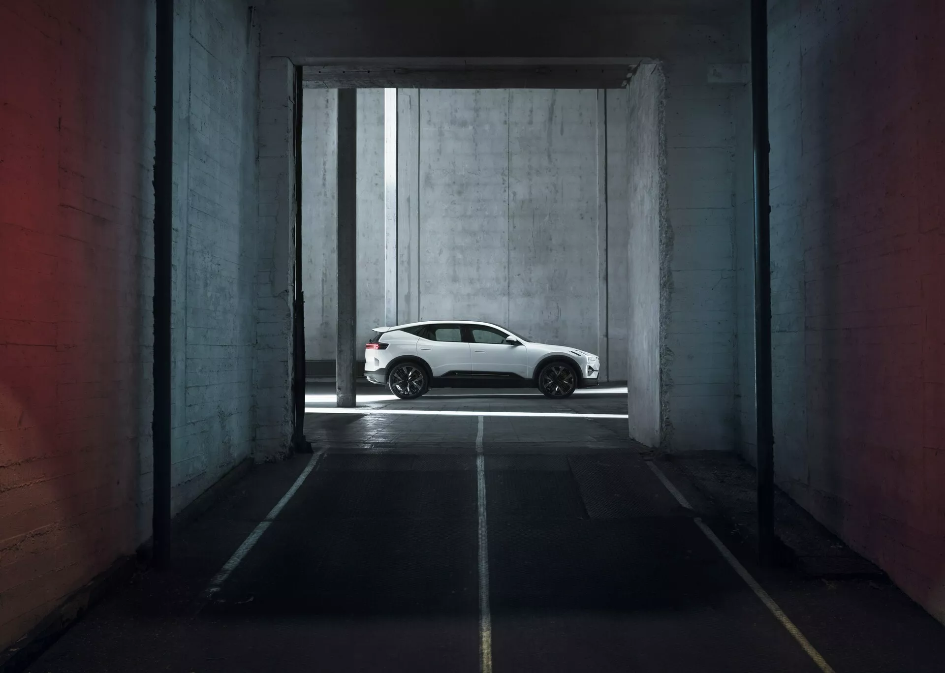 Polestar 3 1ª generazione del 2023 in bianco, che mostra il suo profilo laterale elegante all'interno di una struttura di parcheggio drammaticamente illuminata, evidenziando cerchi in lega sportivi, linee del corpo lisce e aerodinamiche, e finestre oscurate per un aspetto moderno di coupé di lusso.