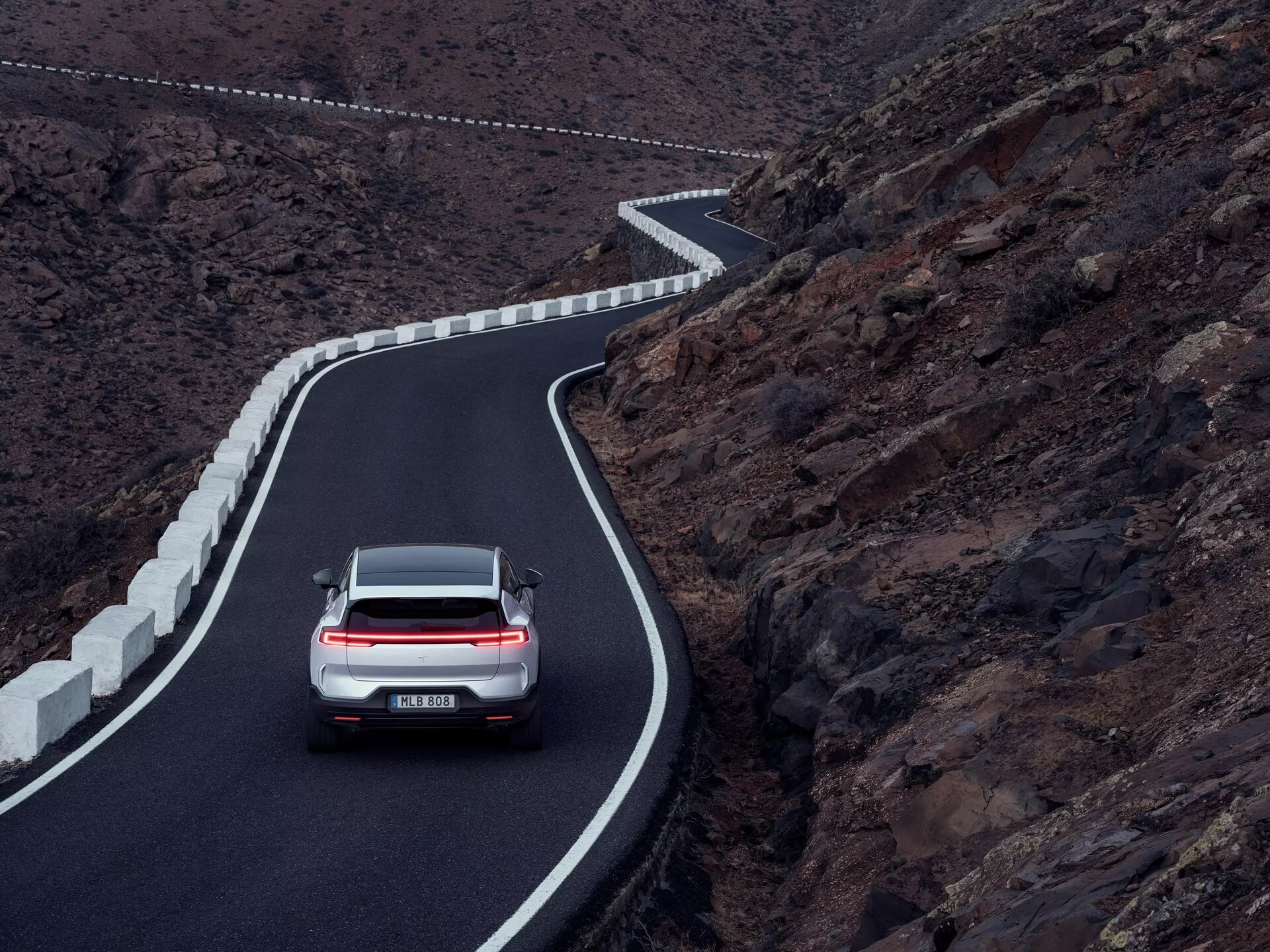 Polestar 3 2023 (1ª generazione) catturata da un punto alto, mostra il suo design posteriore lungo una strada di montagna tortuosa con luci posteriori LED attivate, sottolineando il design automobilistico e le funzionalità tecnologiche avanzate contro uno sfondo montuoso drammatico
