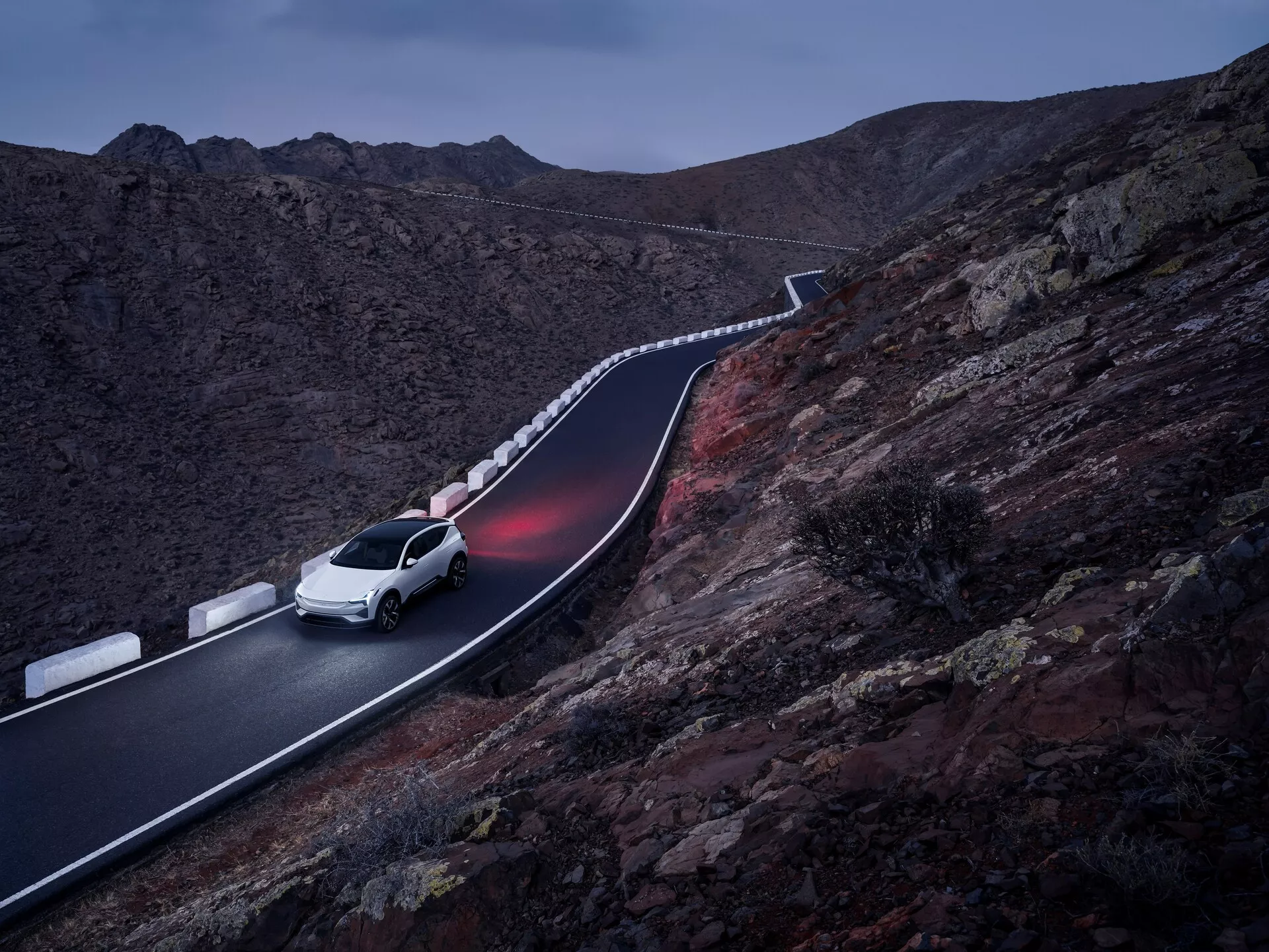 SUV Polestar 3 del 2023 di colore bianco, che guida su una strada montana tortuosa al crepuscolo, mostrando un design elegante e un'illuminazione avanzata