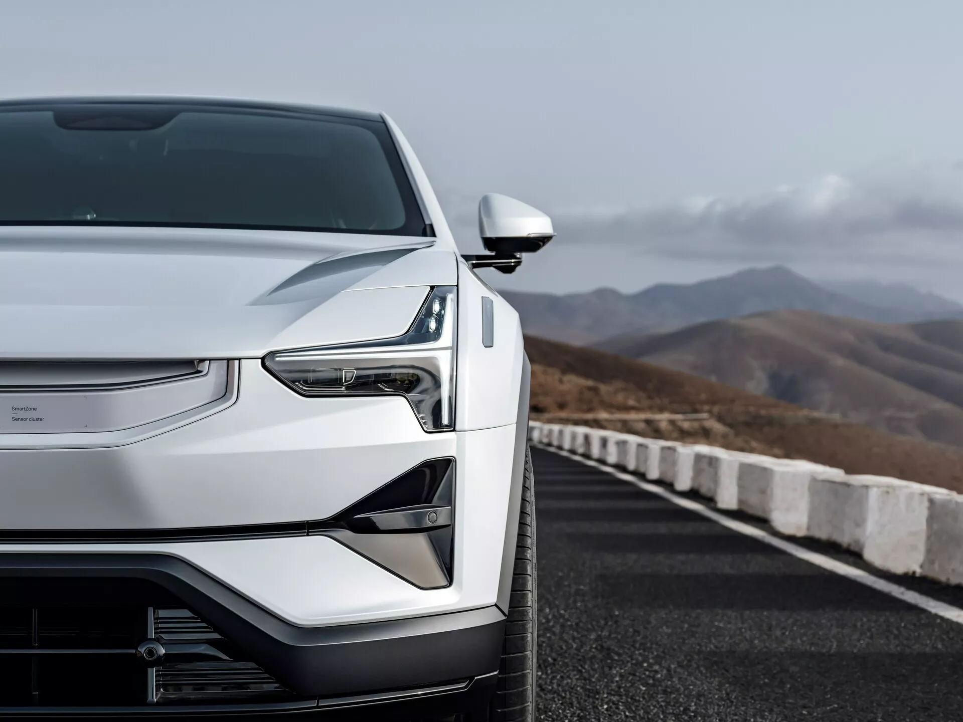 Vista anteriore-sinistra del Polestar 3 del 2023 che mette in mostra il cofano aerodinamico, la griglia chiusa della tecnologia del veicolo elettrico, i fari LED e lo specchietto laterale elegante su una strada con sfondo montagnoso