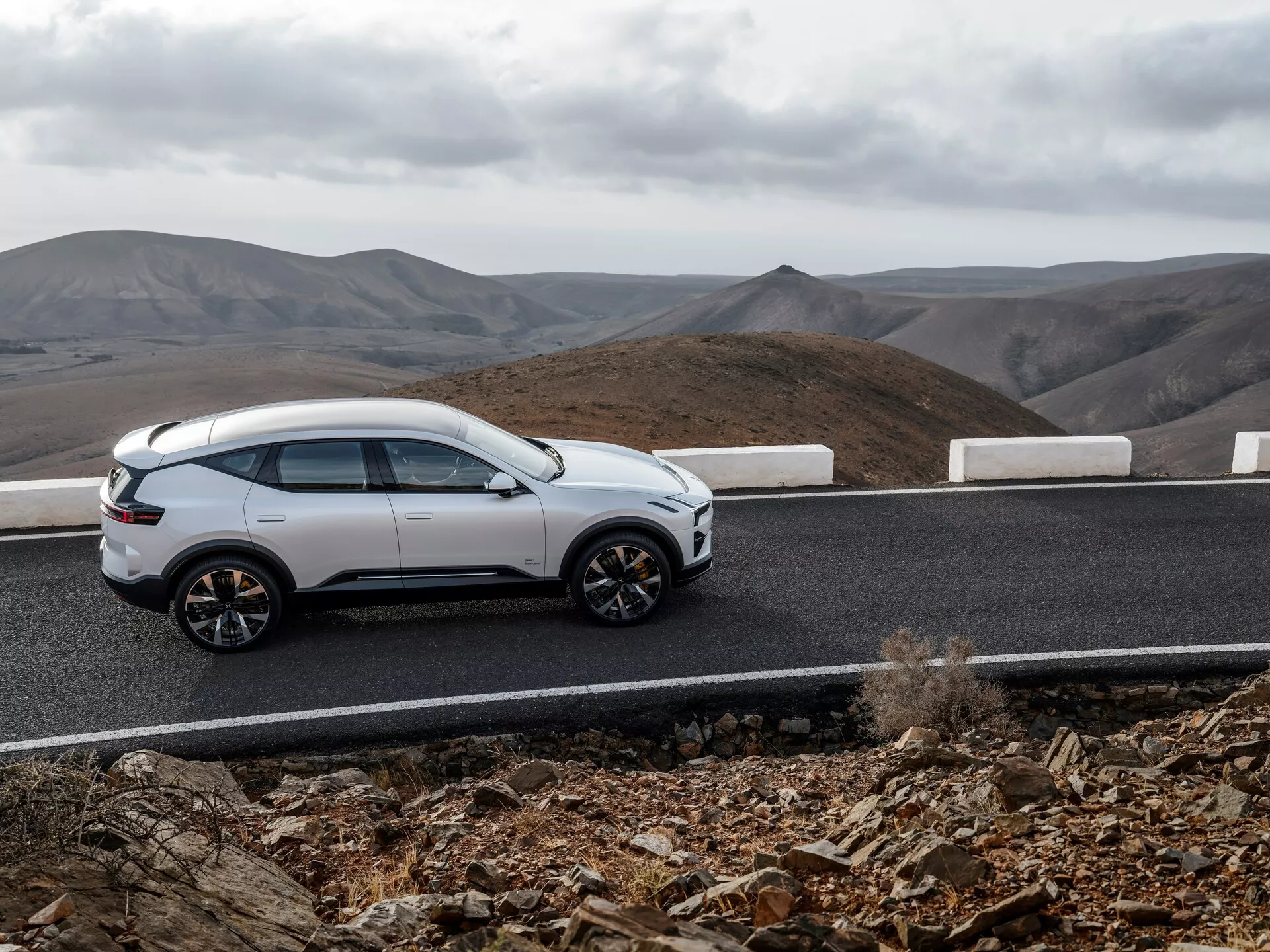 SUV bianca Polestar 3 del 2023 su strada asfaltata curva con bordo bianco contro paesaggio collinare nuvoloso, mostrando profilo laterale, design elegante, ruote a più razze e caratteristiche moderne come maniglie a scomparsa e tetto panoramico, suggerendo lusso e capacità fuoristrada