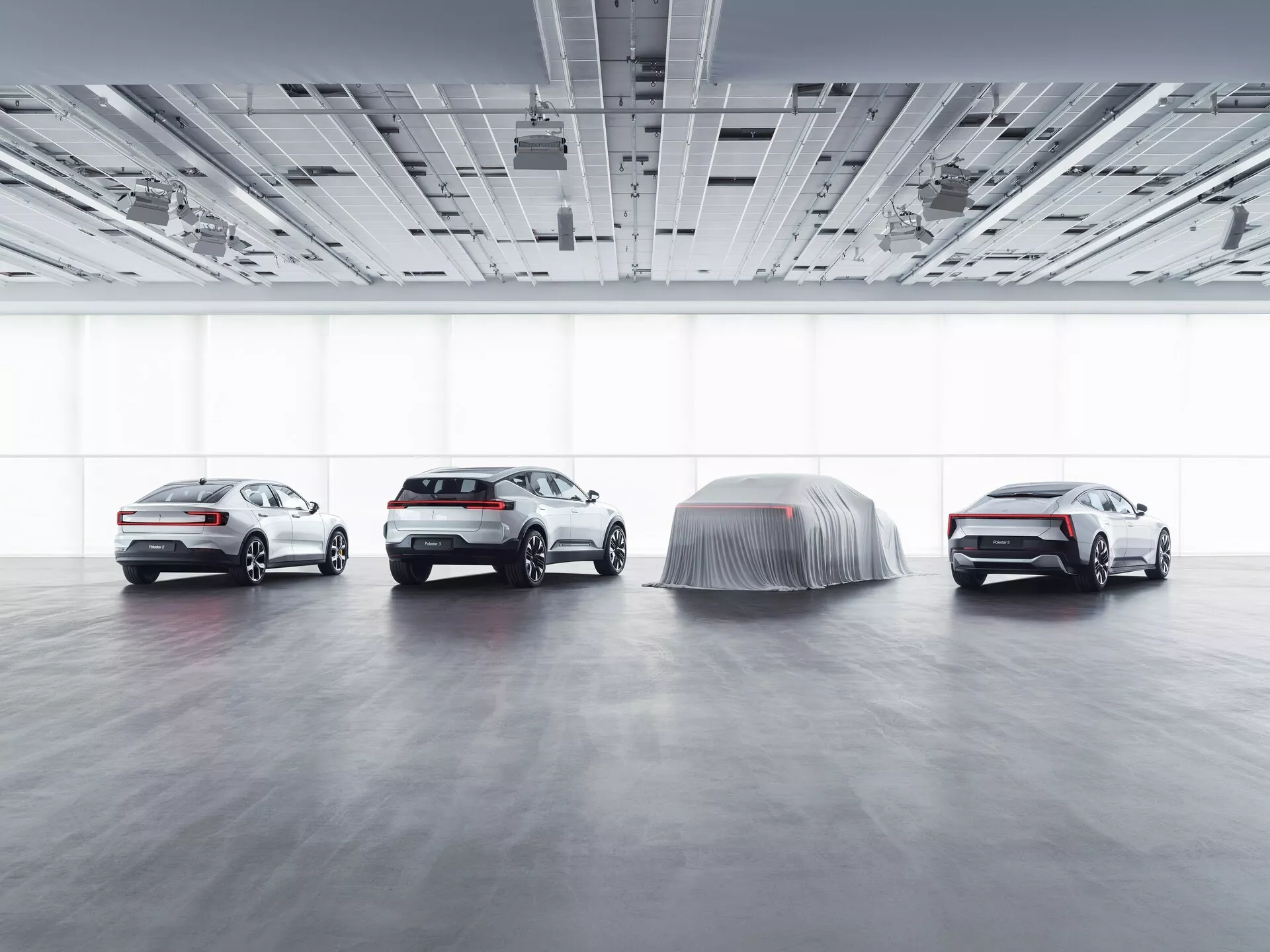 Lineup Polestar 3 2023 in ambientazione showroom con illuminazione diffusa, mostrando viste laterali e frontali di tre auto visibili e un modello coperto che suggerisce una rivelazione, illustrando caratteristiche tecnologiche avanzate