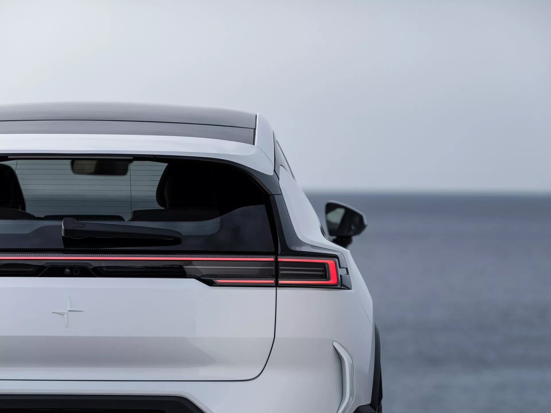 Polestar 3 1ª generazione 2023 che mostra gli elementi di design posteriore, inclusa la striscia di luci posteriori LED orizzontale, finestrino posteriore oscurato con sbrinatore, spoiler integrato nel bagagliaio e badge premium, su uno sfondo sereno con acqua aperta.