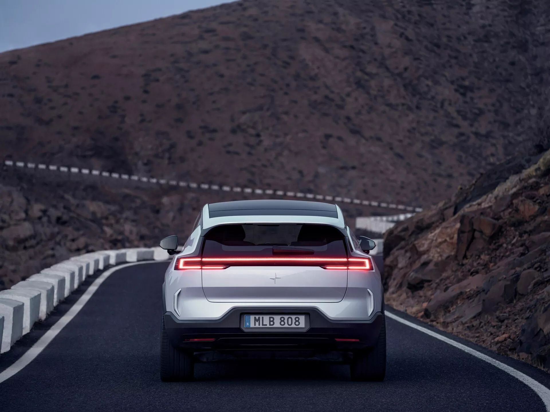 Vista posteriore di una Polestar 3 2023 catturata al crepuscolo su una strada montagnosa, mostrando la sua barra luminosa a tutta larghezza, sensori di parcheggio e assenza di scarichi visibili, indicando un veicolo elettrico o ibrido di lusso