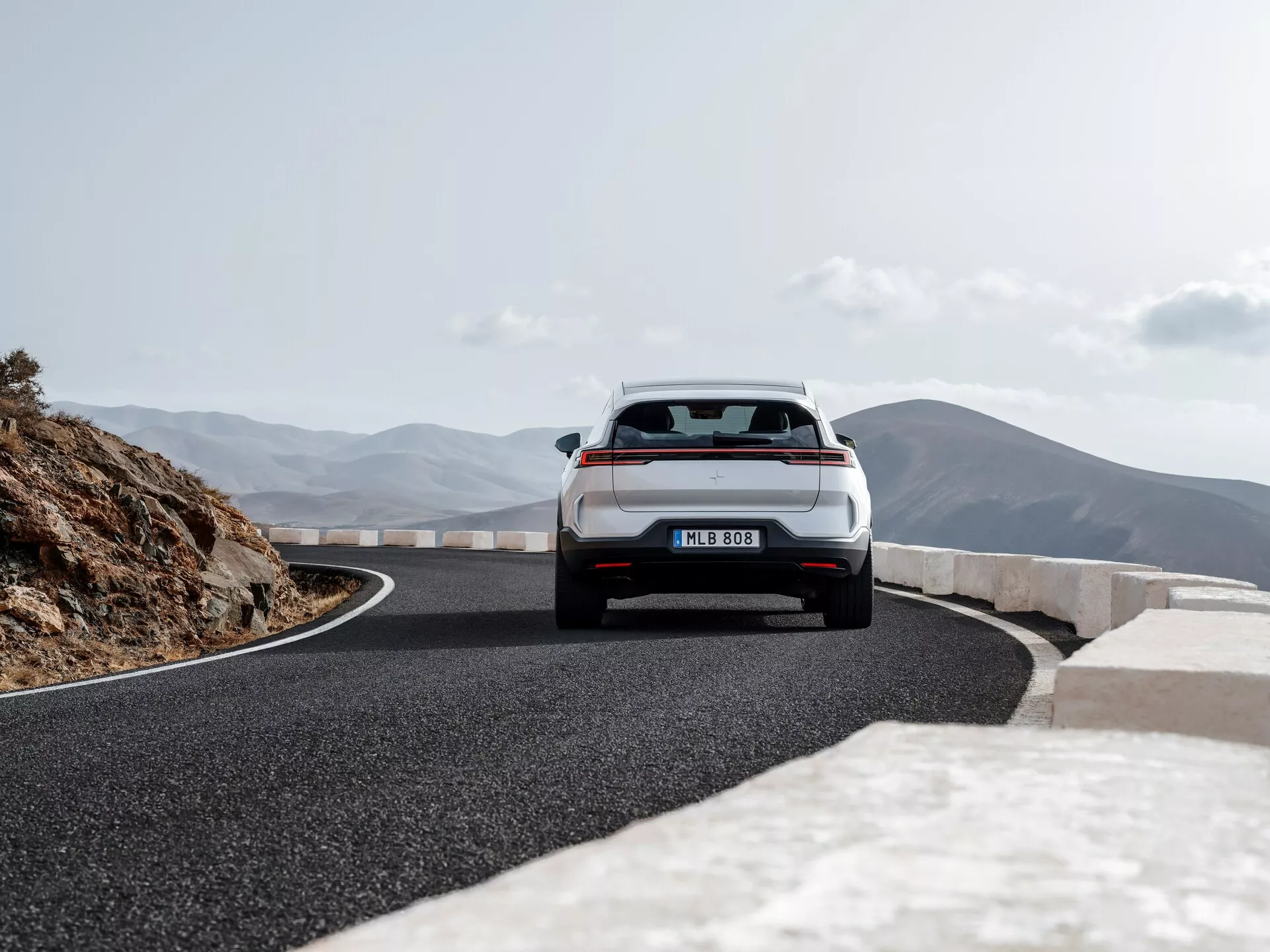 Polestar 3 prima generazione del 2023 catturata da una prospettiva posteriore che mostra il suo retro completo inclusi bagagliaio, paraurti posteriore, luci posteriori e parte del tetto posteriore su una strada di montagna panoramica, evidenziando il suo design hatchback o SUV crossover con luci posteriori LED, sistema di scarico doppio e tergicristallo posteriore per una maggiore sicurezza e prestazioni