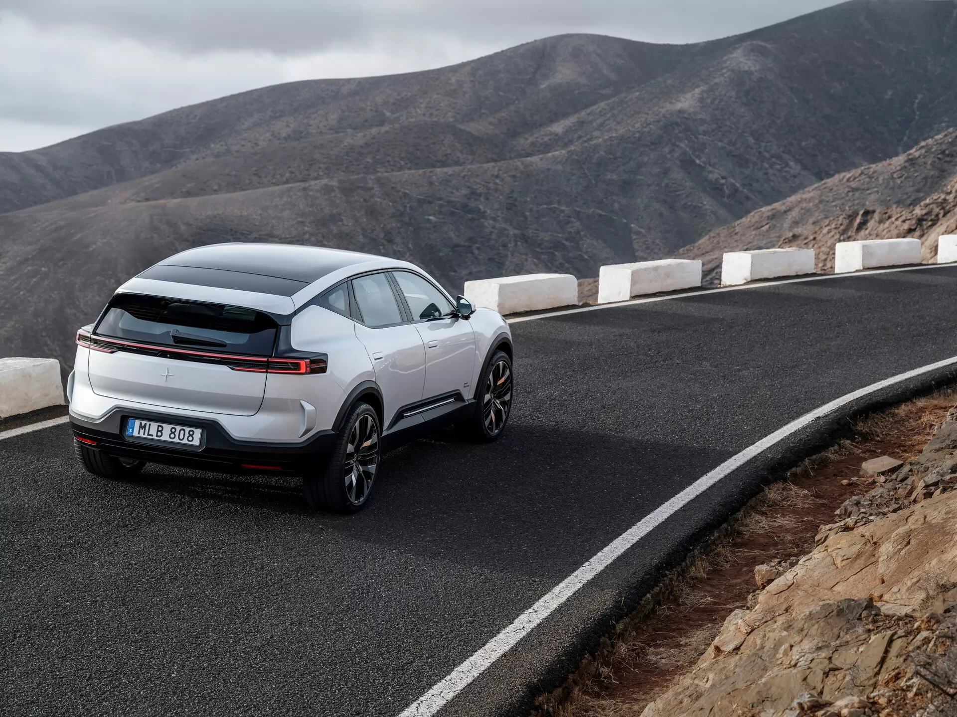SUV crossover bianco Polestar 3 prima generazione 2023 che mostra un design elegante e sportivo con grandi ruote in lega e distintive luci posteriori LED, parcheggiato su una strada montana tortuosa, vista posteriore da tre quarti che enfatizza le sue linee moderne e funzionalità tecnologiche.