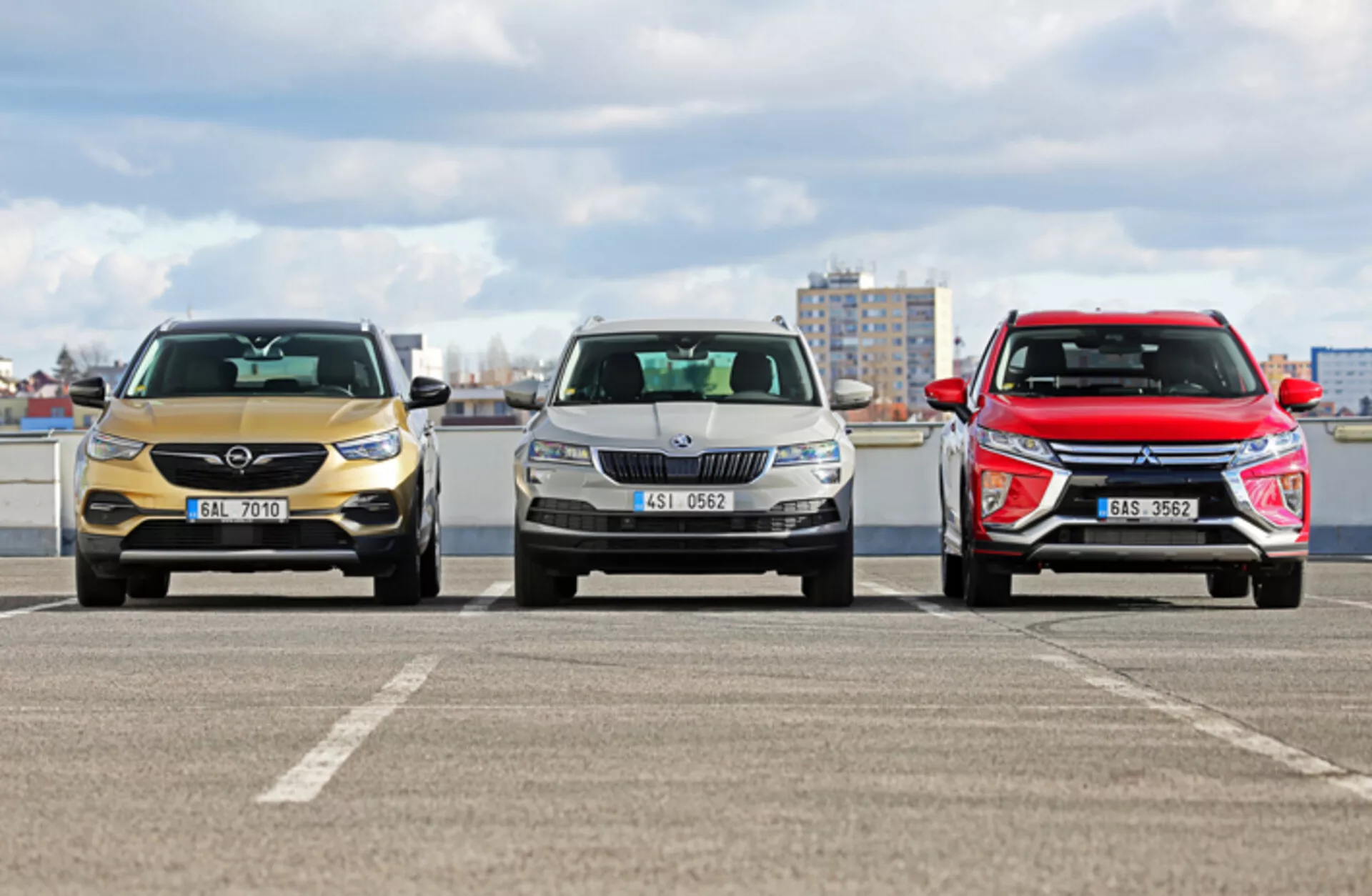 Vista frontale di tre auto parcheggiate una accanto all'altra che mostrano caratteristiche di design tra cui griglie, fari LED e paraurti anteriori; un veicolo oro con accenti cromati a sinistra, un veicolo argento con un design conservativo al centro, e un modello rosso sportivo o SUV a destra, fotografato da un angolo basso su un parcheggio sul tetto.