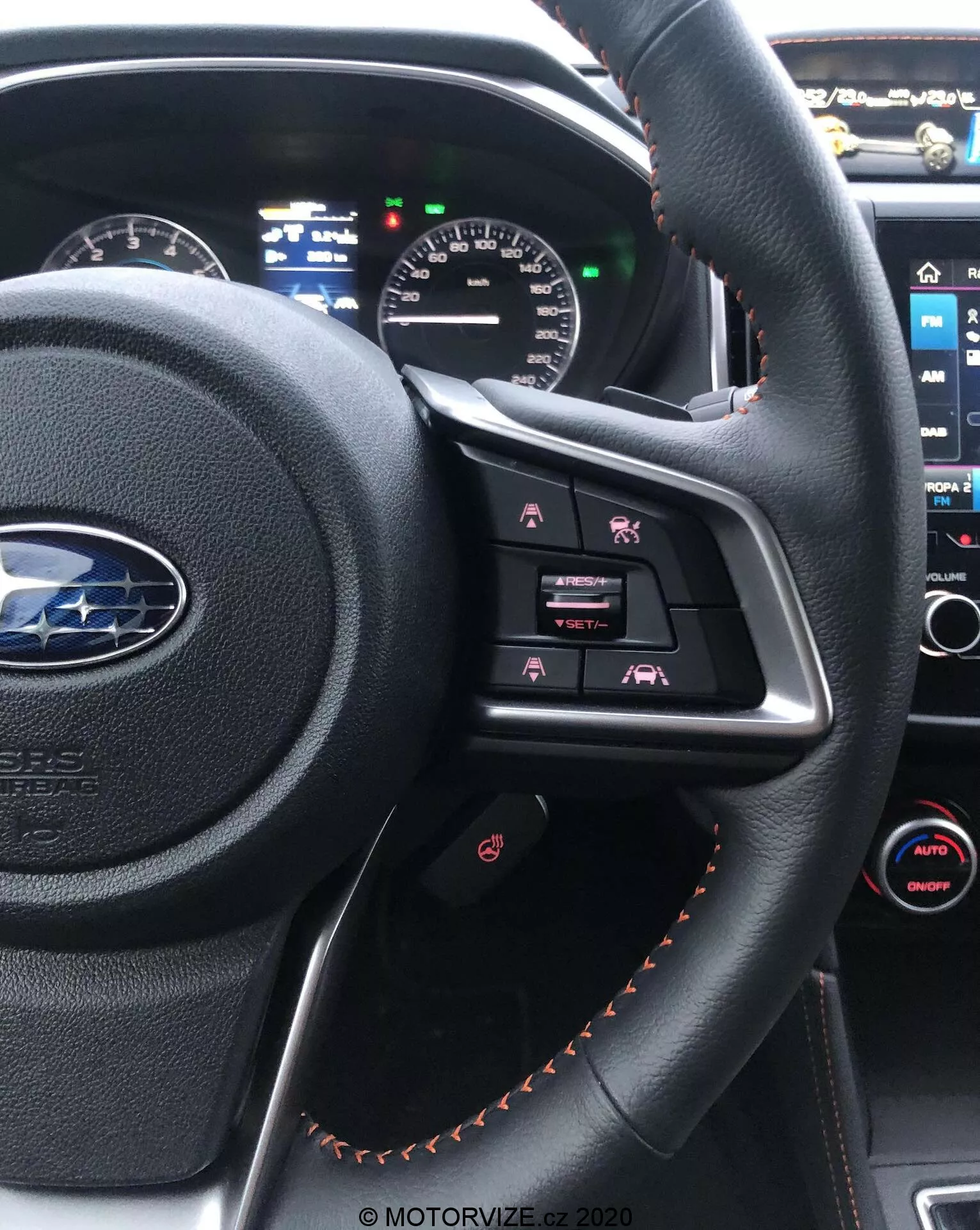 Vista interna di una Subaru XV 2018 dal sedile del conducente, evidenziando il volante rivestito in pelle nera con cuciture arancioni, con pulsanti di controllo multimediali e un quadro strumenti parzialmente visibile con display analogici e digitali. Sullo sfondo è visibile la console centrale con il suo touchscreen di infotainment e il controllo del clima automatico a doppia zona.