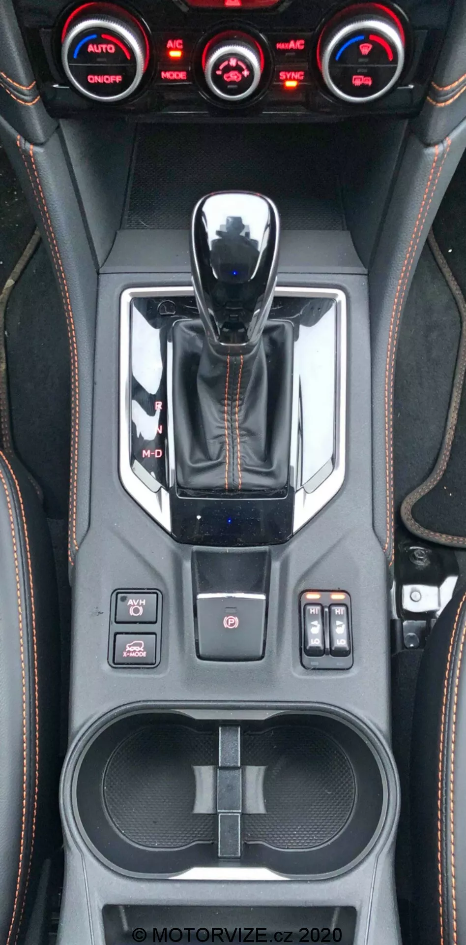 Vista ravvicinata verticale della console centrale della Subaru XV 2018, che mostra la leva del cambio con finitura metallica, il selettore delle modalità di guida, vari pulsanti di controllo tra cui controllo della trazione, sensori di parcheggio e impostazioni delle sospensioni, sistema di controllo del clima a tre zone, dettagli cuciti a mano in similpelle, doppio portabicchieri, vano portaoggetti con porte USB e filigrana visibile MOTORVIZE.cz.