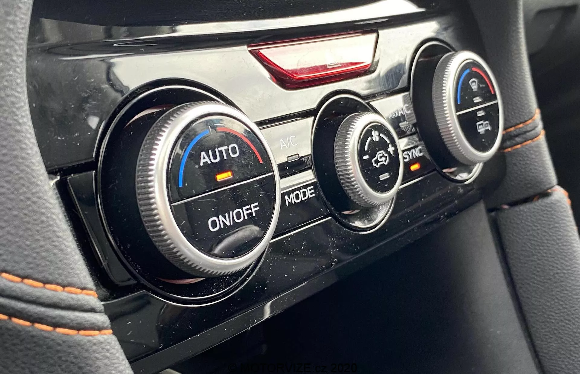 Vista ravvicinata delle manopole di controllo del clima all'interno della Subaru XV 2018. Scattata dalla prospettiva del conducente, mostra tre manopole circolari: quella a sinistra in modalità 'AUTO' per la velocità della ventola, quella centrale per la selezione della modalità 'ON/OFF' e quella di destra per il controllo della temperatura del passeggero con modalità 'SYNC'. Sopra la manopola centrale c'è un pulsante di emergenza rosso. L'immagine mette in evidenza accenti cromati sulle manopole e cuciture rosse sotto di esse.