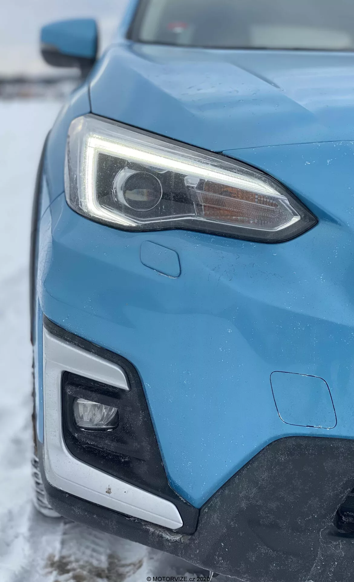 Vista ravvicinata del lato anteriore sinistro di una Subaru XV 1.6i Trend Lineartronic blu del 2018, catturata da un'angolazione obliqua che si concentra sul faro e su una parte della griglia e del paraurti anteriore. Il faro anteriore sinistro prominente presenta una lente del proiettore indicativa di sistemi HID o LED e luci diurne a LED integrate. L'immagine mostra anche una parte del paraurti anteriore che probabilmente ospita un'illuminazione aggiuntiva. La carrozzeria dell'auto mostra linee nitide e forme aerodinamiche, con uno sfondo invernale con una leggera spolverata di neve.