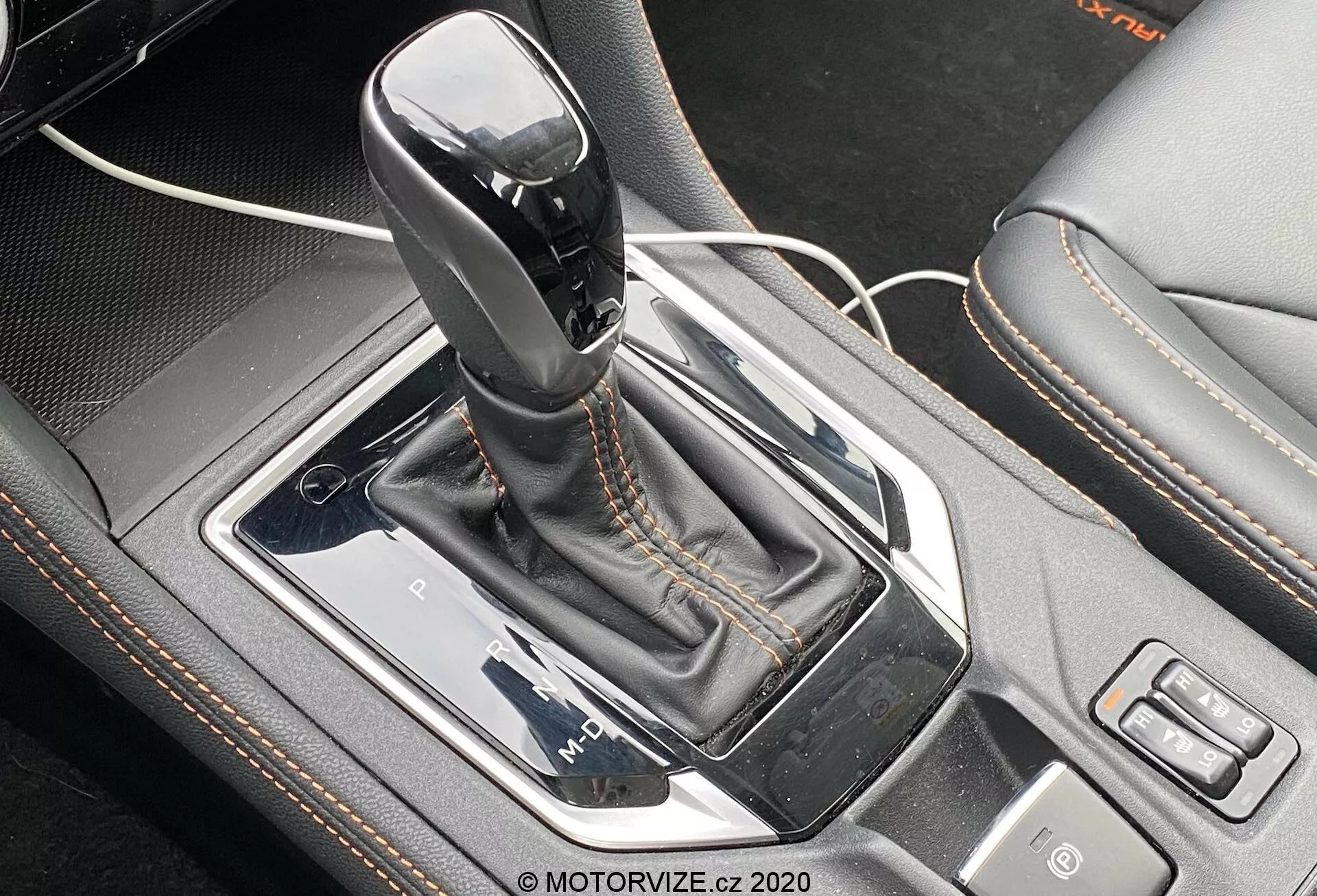 Vista ravvicinata della leva del cambio automatico Lineartronic della Subaru XV 2018 1.6i Trend. La leva del cambio, in posizione di parcheggio e con una parte superiore nera lucida con accento argento, è posizionata centralmente nella console centrale dell'auto. È circondata da un pannello argento, racchiusa in una cornice metallica nera lucida con un soffietto in finta pelle con cuciture a contrasto. A sinistra, un cavo USB indica una porta vicina, mentre a destra sono visibili i pulsanti per la selezione del modo di guida e un'altra funzione. La prospettiva dall'alto e leggermente di fronte enfatizza i dettagli di design complessi e le caratteristiche tecnologiche dell'area della leva del cambio.