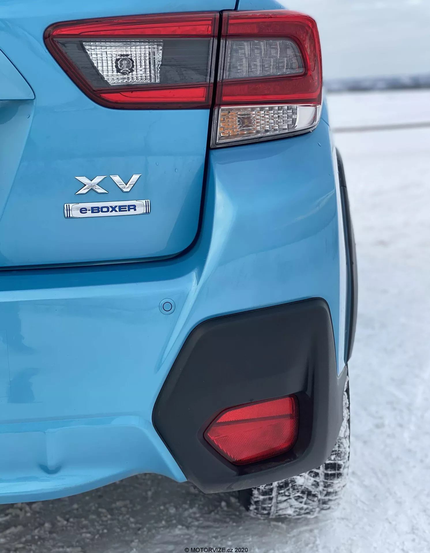 Una ripresa ravvicinata e angolata posteriore di un modello Subaru XV 2018 blu, focalizzata sull'angolo posteriore sinistro dalla luce posteriore alla ruota posteriore. Le caratteristiche principali includono un complesso cluster di luci posteriori, badge 'XV' e 'e-BOXER' che indicano tecnologia ibrida, vernice blu metallizzata lucida con modanatura ad arco ruota in plastica nera, un inserto in plastica nera sul paraurti posteriore che riflette un terreno innevato che suggerisce un clima freddo e un pneumatico posteriore parzialmente visibile con battistrada ricoperto di neve. La messa a fuoco è nitida sull'angolo posteriore, mentre lo sfondo è leggermente sfocato a causa della profondità di campo.