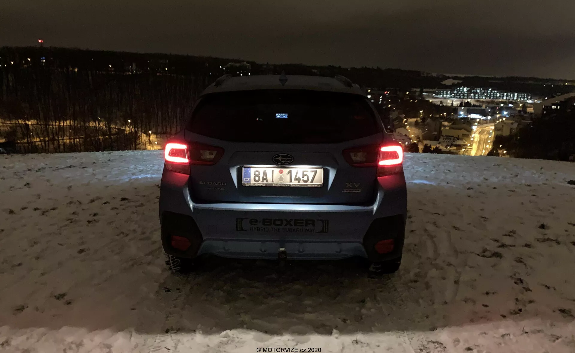 Vista posteriore della Subaru XV 2018 (subaru-xv-1-6i-trend-lineartronic) catturata di notte con le luci della città sullo sfondo. L'auto è in una zona elevata, mostrando le luci posteriori illuminate, il parabrezza posteriore, il badge del produttore, l'indicazione del modello e-BOXER, la targa, il paraurti posteriore e la telecamera o il sensore di parcheggio. Le superfici riflettenti e la neve a terra suggeriscono condizioni invernali. Il design moderno del veicolo comprende un piccolo spoiler, un tergicristallo posteriore minimalista e un paraurti bicolore, mettendo in risalto gli elementi di illuminazione automobilistica e di design del marchio.