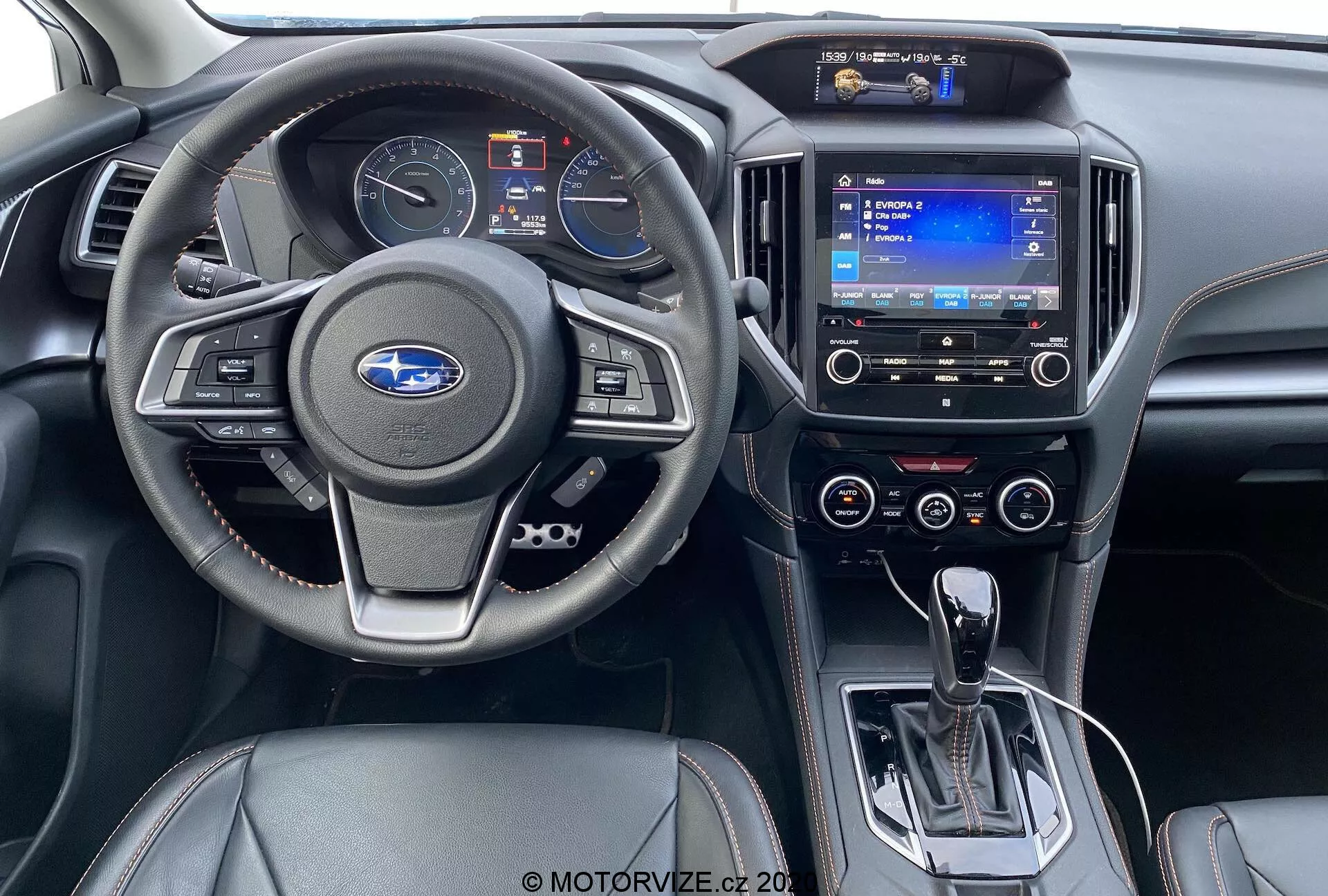 Vista dell'interno dal sedile posteriore di una Subaru XV 1.6i Trend Lineartronic 2018. Presenta un moderno volante con pulsanti per il controllo audio e della velocità, un quadro strumenti parzialmente visibile, un sistema di infotainment touchscreen con navigazione, comandi del climatizzatore a doppia zona, leva del cambio automatico in posizione di parcheggio, interruttore del freno di stazionamento elettronico, comandi del riscaldamento dei sedili e interni in pelle nera bicolore con accenti argentati.