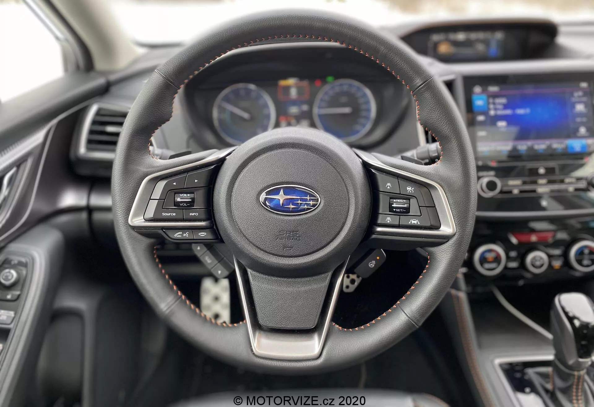 Prospettiva interna di una Subaru XV 2018 (Subaru-xv-1-6i-trend-lineartronic) con messa a fuoco sul volante e sulla plancia. Il volante a tre razze, rivestito in pelle con cuciture visibili, presenta pulsanti per il controllo multimediale e del cruise control. Dietro di esso, la plancia mostra due indicatori analogici separati da uno schermo digitale di informazioni per il conducente. Sullo sfondo, leggermente sfocato, è visibile il sistema di infotainment touchscreen e i comandi del clima. Le cuciture rosse sul volante e sul rivestimento della plancia sottolineano un livello di finitura sportivo o premium. Scattata dal punto di vista del conducente, questa immagine moderna e ben curata enfatizza la tecnologia e il design automobilistico.