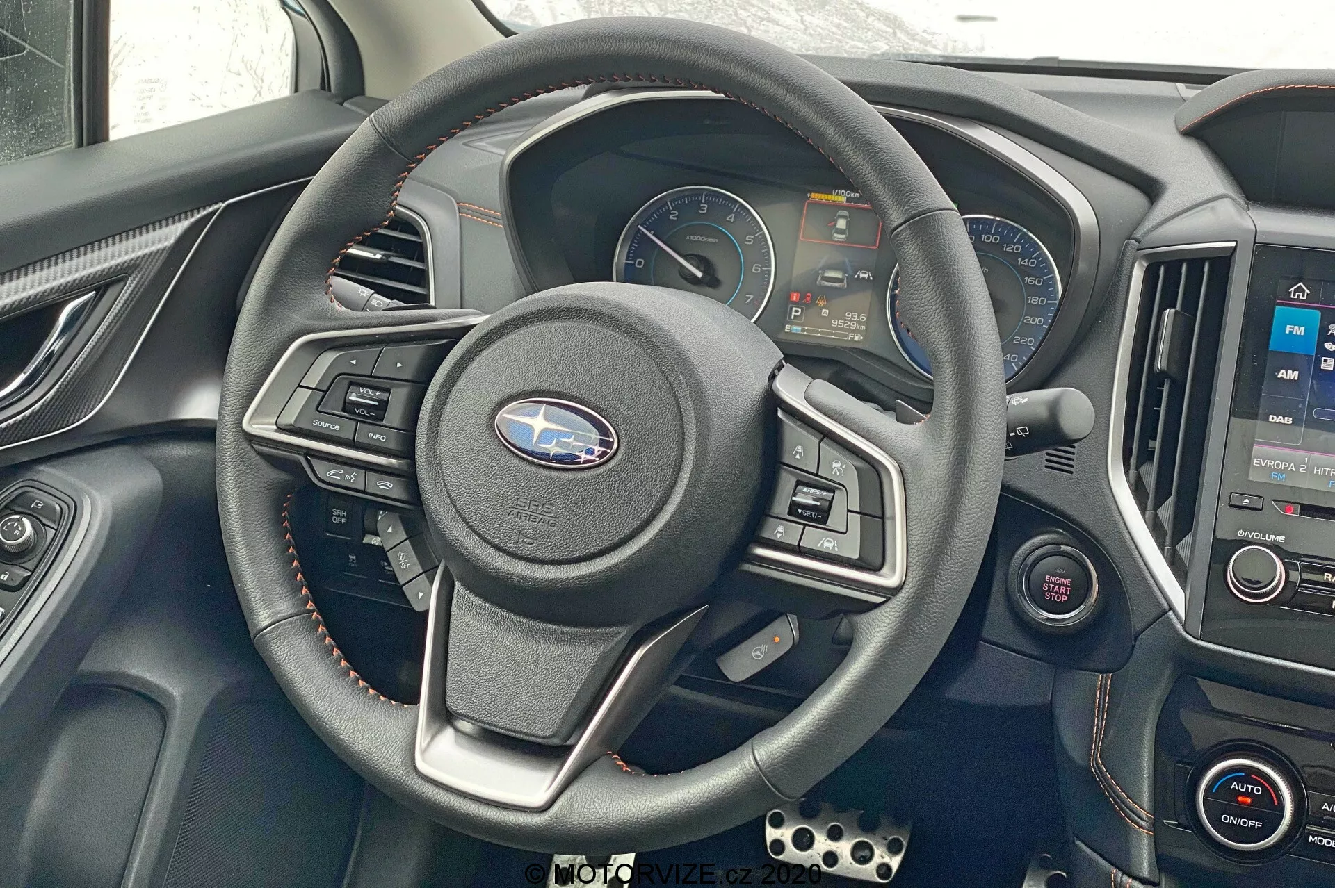 Scatto interno della Subaru XV 2018, focalizzato sul volante e sull'area del cruscotto. Volante a tre razze centrale con comandi integrati, quadro strumenti digitale che mostra tachimetro, contagiri e schermi informativi. Consolle centrale con sistema di infotainment che mostra una mappa e comandi del clima sotto. Accenti metallici, volante rivestito in pelle, dettagli rossi sulle bocchette. Ulteriori pulsanti di controllo a sinistra, pedali in stile sportivo visibili in basso a sinistra. Inquadratura frontale, leggermente verso il basso, che mostra comandi e tecnologia del conducente.